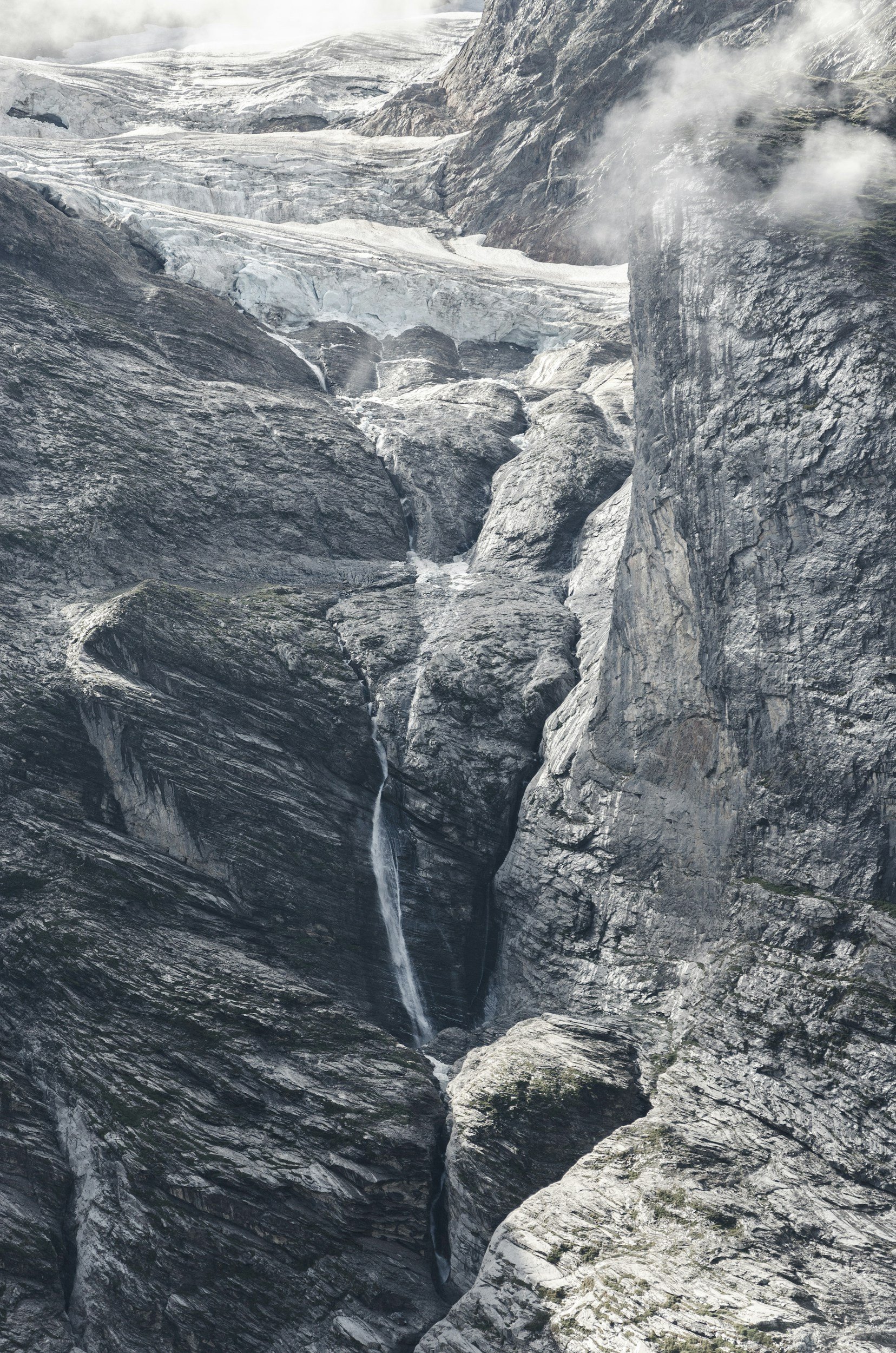 A rugged mountain landscape with a glacier at the top, steep cliffs, and a thin waterfall cascading down the rocks.