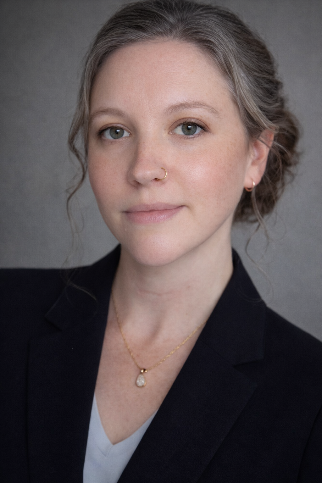 Headshot of a woman with light skin, shoulder-length curly hair, and blue eyes, wearing a black blazer, a white top, gold jewelry, and a nose ring, against a plain gray background.