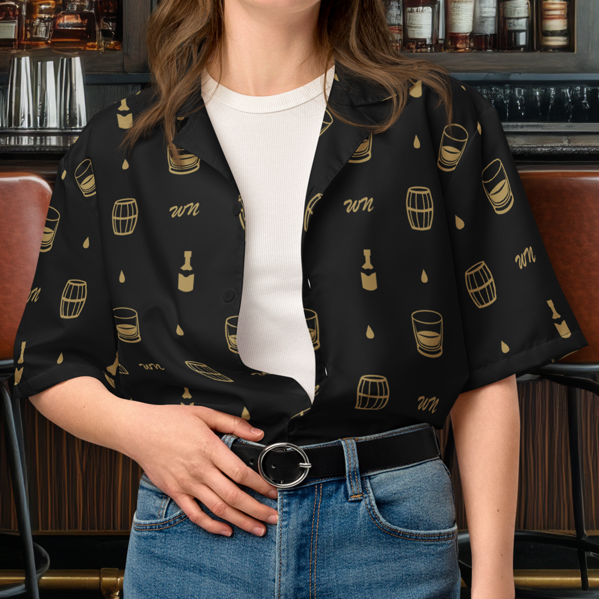 Close-up of a woman wearing a black button-up shirt with beer-themed patterns, a white undershirt, and blue jeans with a black belt, sitting in a bar.
