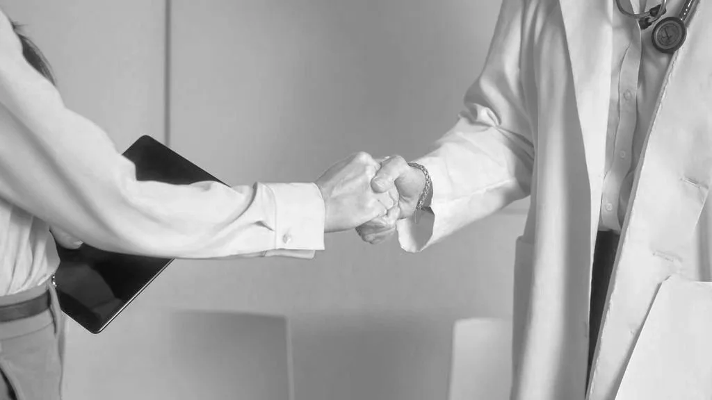 A doctor and a patient shaking hands in a hospital or medical office setting, both dressed in professional attire, with the doctor wearing a white coat and stethoscope.