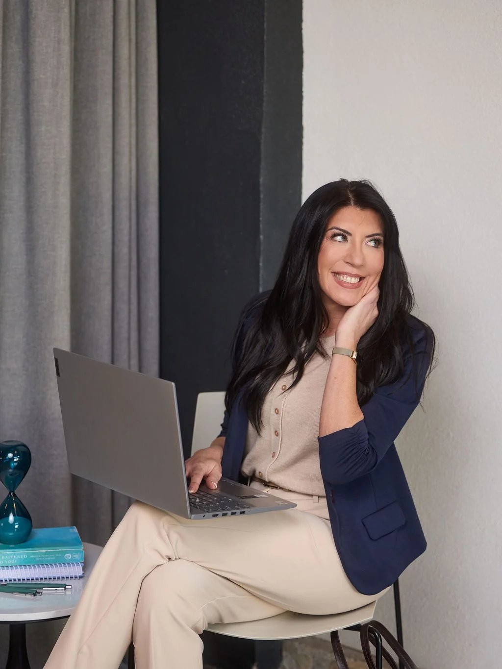 A woman with long dark hair smiling, sitting on a chair with a laptop on her lap, near a side table with books, a pen, and a decorative hourglass, against a background of a white wall and gray curtains.