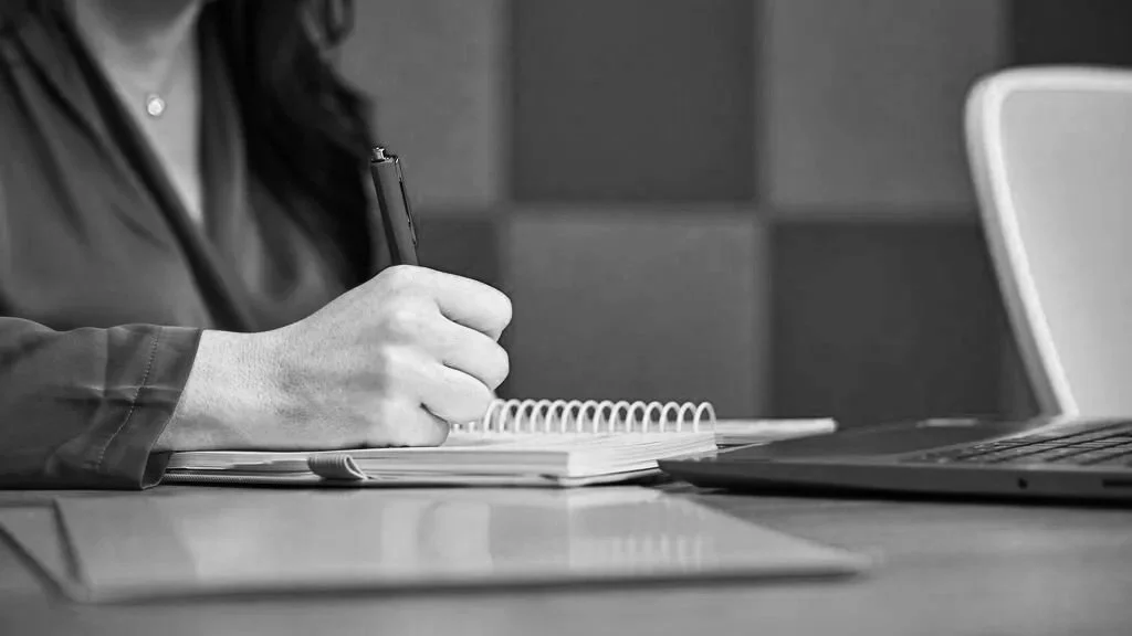 Person taking notes at a desk with a laptop open beside them in a modern office setting
