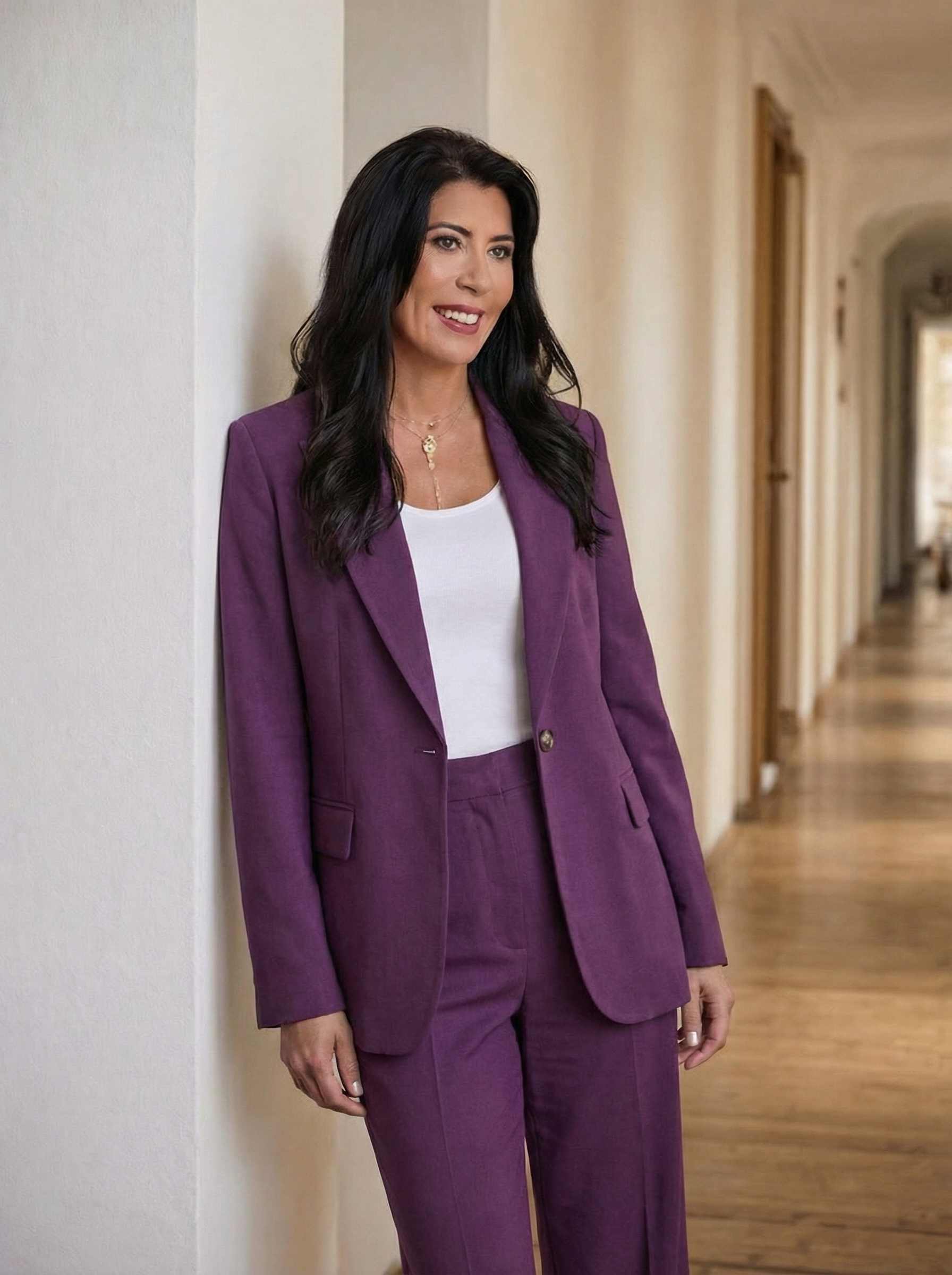 A woman with long black hair, wearing a purple suit jacket and pants with a white top, standing in a hallway.