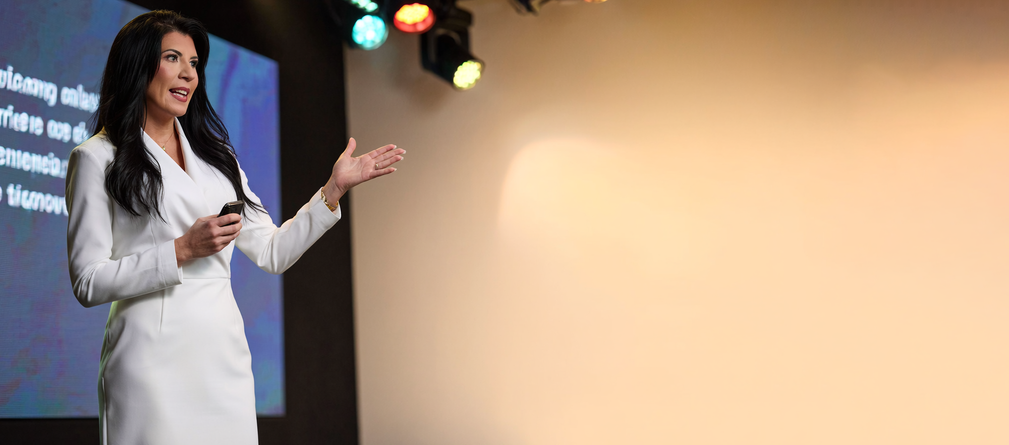 A woman dressed in a white blazer and skirt, speaking at a presentation with a microphone in her hand, gesturing with her other hand, with a colorful stage light setup behind her.