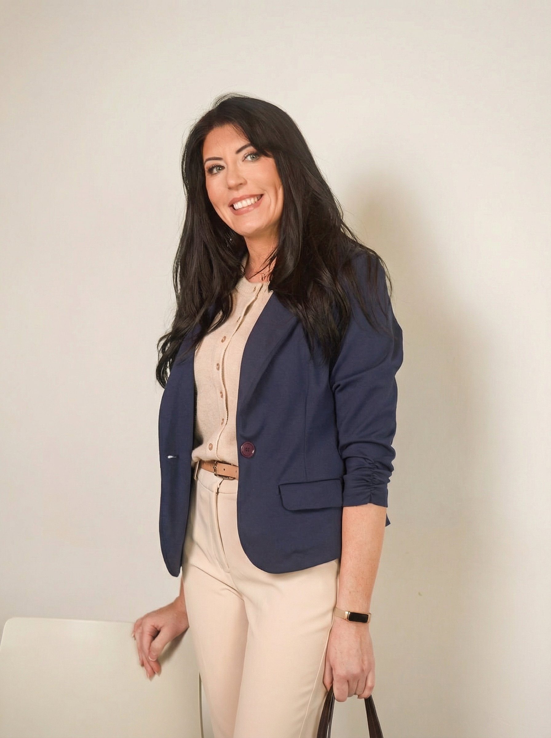 Woman with long dark hair smiling, dressed in a beige blouse and cream pants, wearing a navy blazer and holding a black handbag against a plain wall.