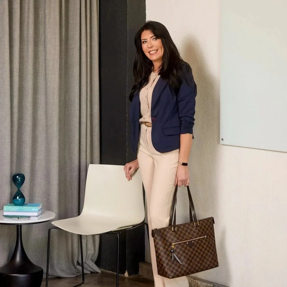 A woman standing indoors, smiling, wearing a dark blazer, beige shirt and trousers, holding a checkered brown handbag.