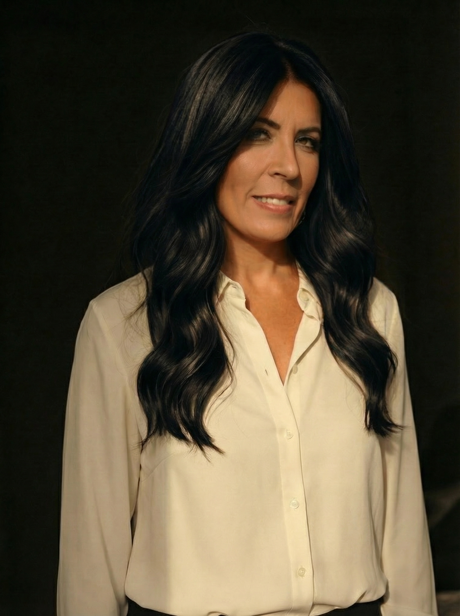 A woman with long, wavy black hair wearing a cream-colored blouse, standing against a dark background.