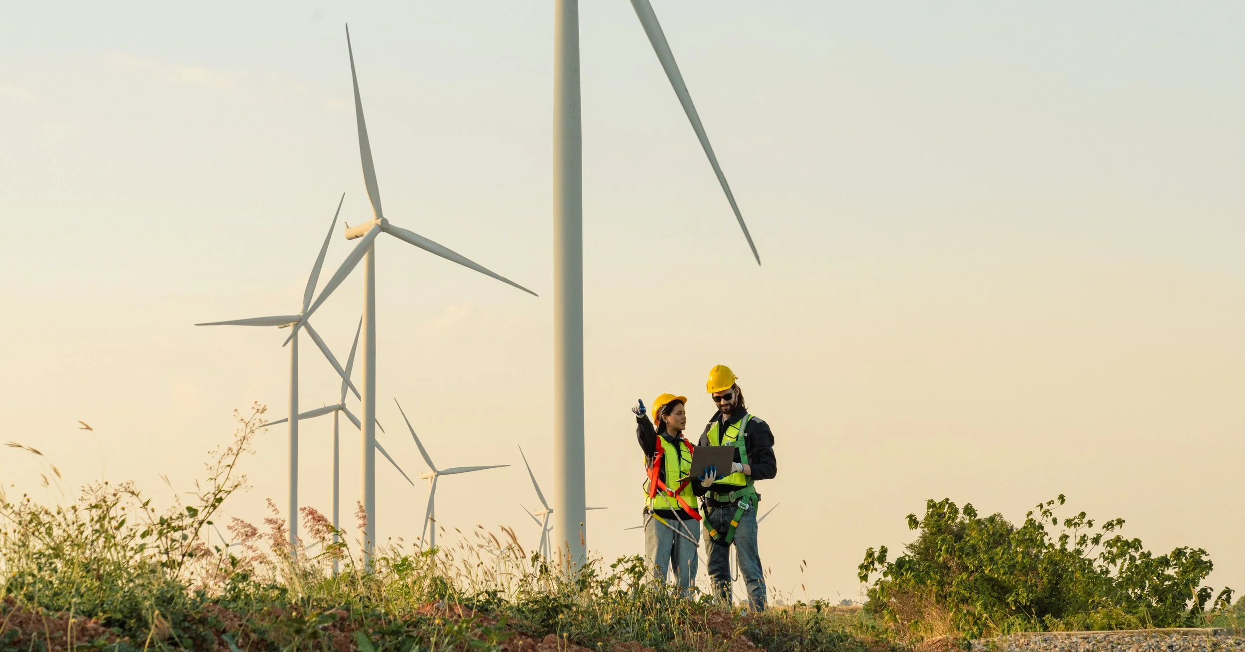 Two wind turbine technicians, wearing yellow hard hats and safety vests, inspecting wind turbines with a laptop in a wind farm during sunset.