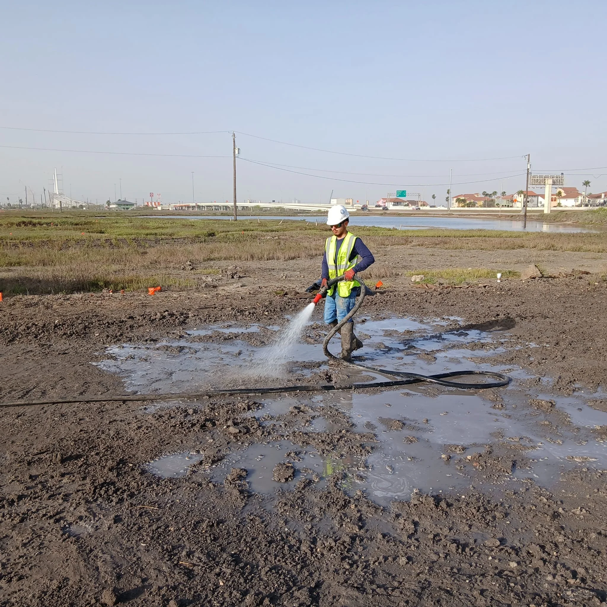 Worker in safety gear using a hose to spray water on muddy ground in an open field with buildings and power lines in the background.