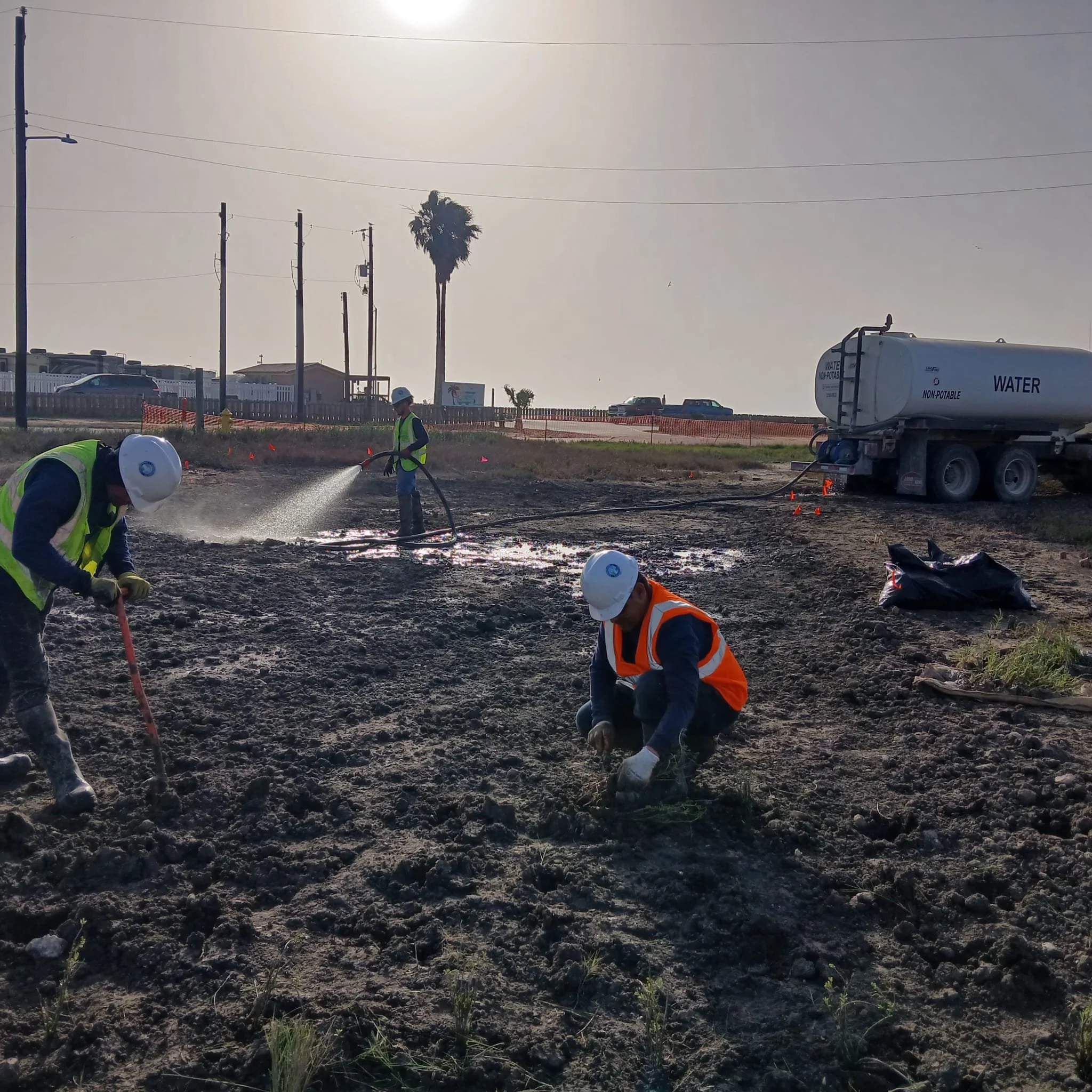 Construction workers applying water to a dirt site, with a water tanker truck nearby, under a sunny sky.