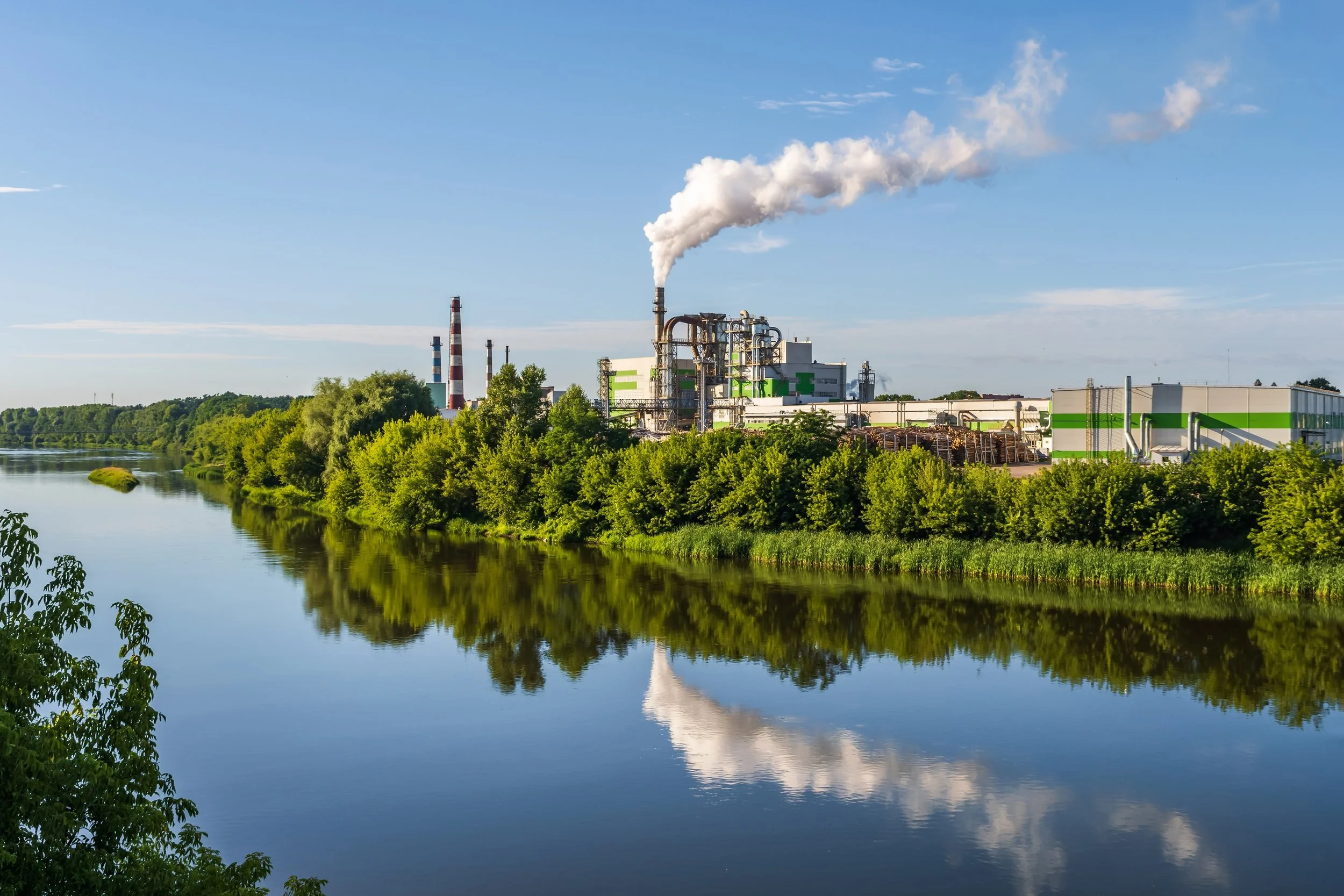 Industrial factory with smoke emissions near a river, lush green trees, and a partly cloudy sky.
