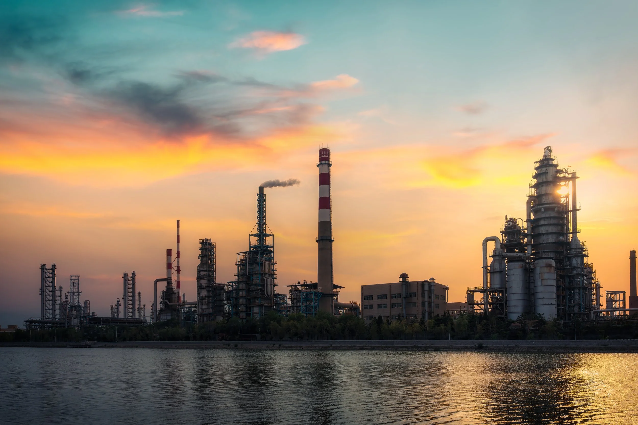 Industrial factory with smokestacks and pipes at sunset, reflecting on a body of water in the foreground.