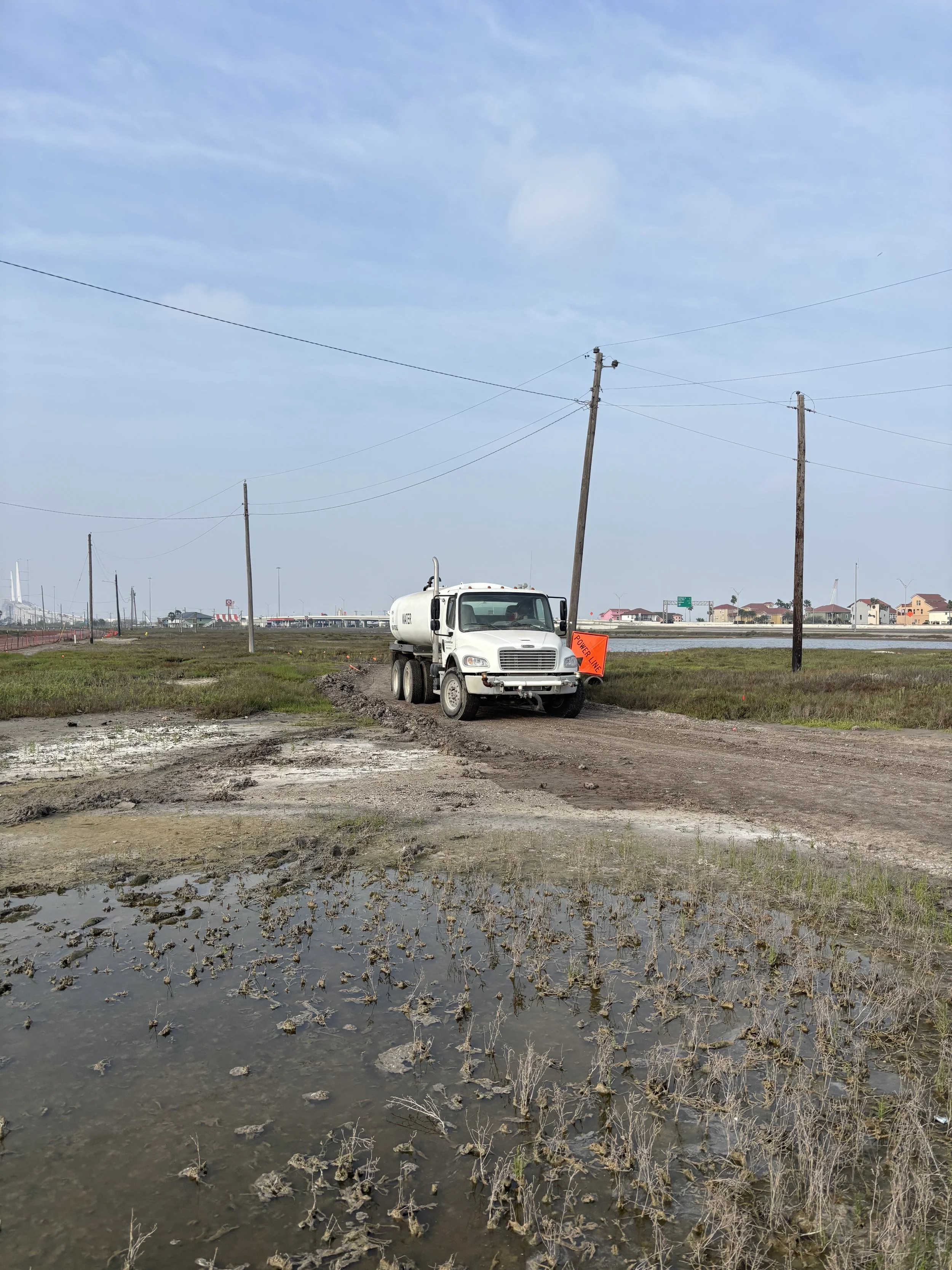 A white utility truck parked on a dirt road with a sign saying 'Power Outage' next to it, and power lines in a rural area with wet ground and a cloudy sky.