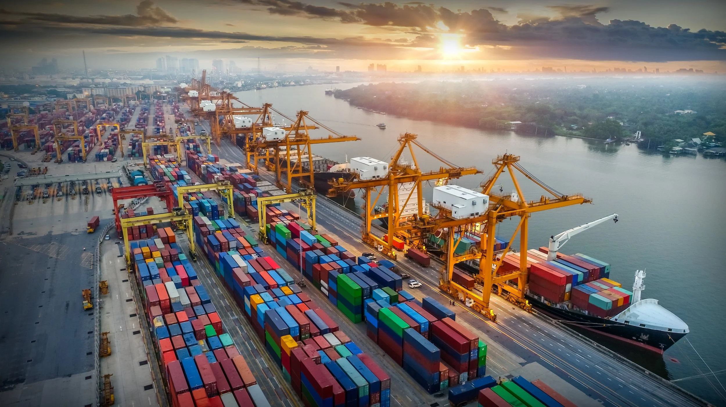 An aerial view of a busy container port with numerous shipping containers, large yellow gantry cranes, a cargo ship docked at the port, and a river with a cityscape in the background during sunset.
