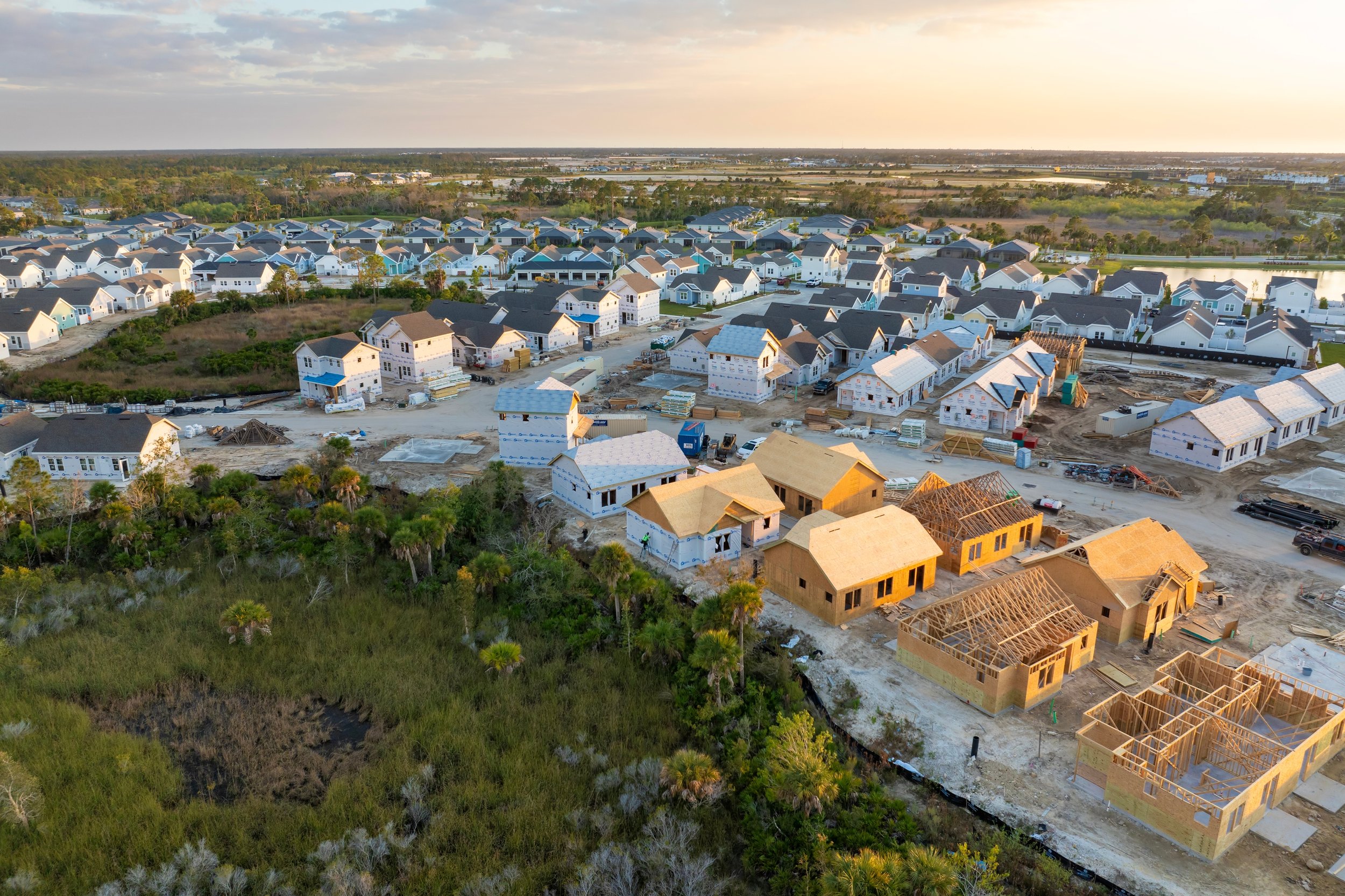 Aerial view of a residential construction site with houses in various stages of building, surrounded by a neighborhood of completed homes and green landscape.