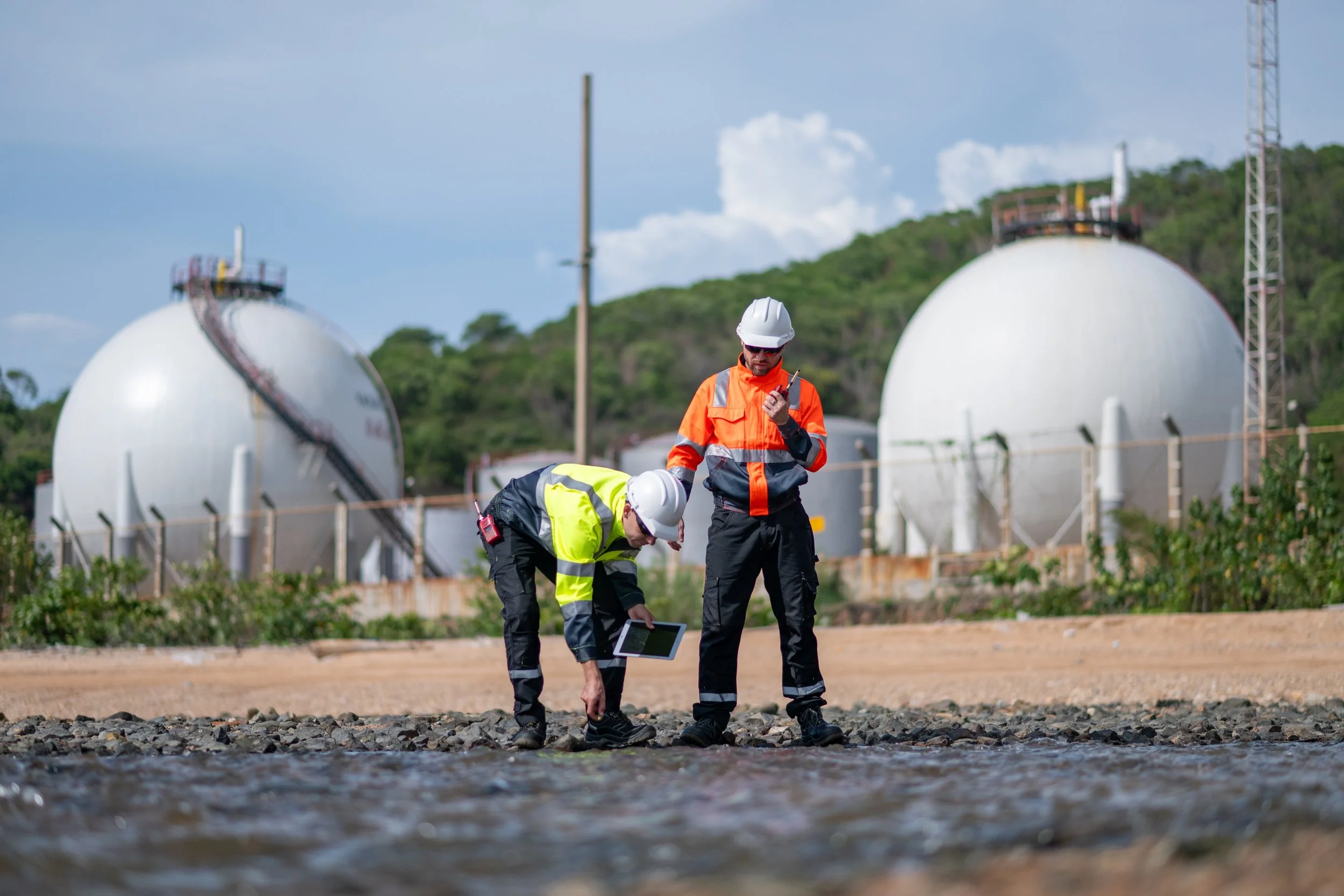 Two workers in safety helmets and reflective clothing inspecting a body of water near industrial tanks.