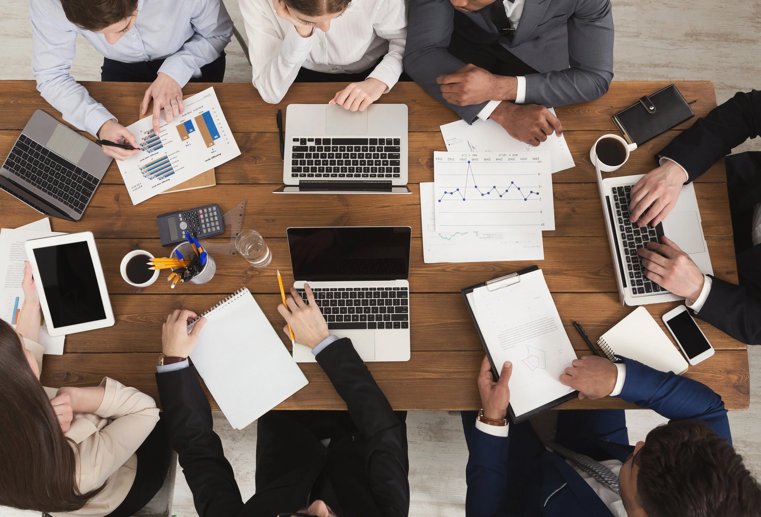 Overhead view of a diverse group of people sitting around a wooden conference table with laptops, tablets, papers, charts, notebooks, pens, and cups of coffee.