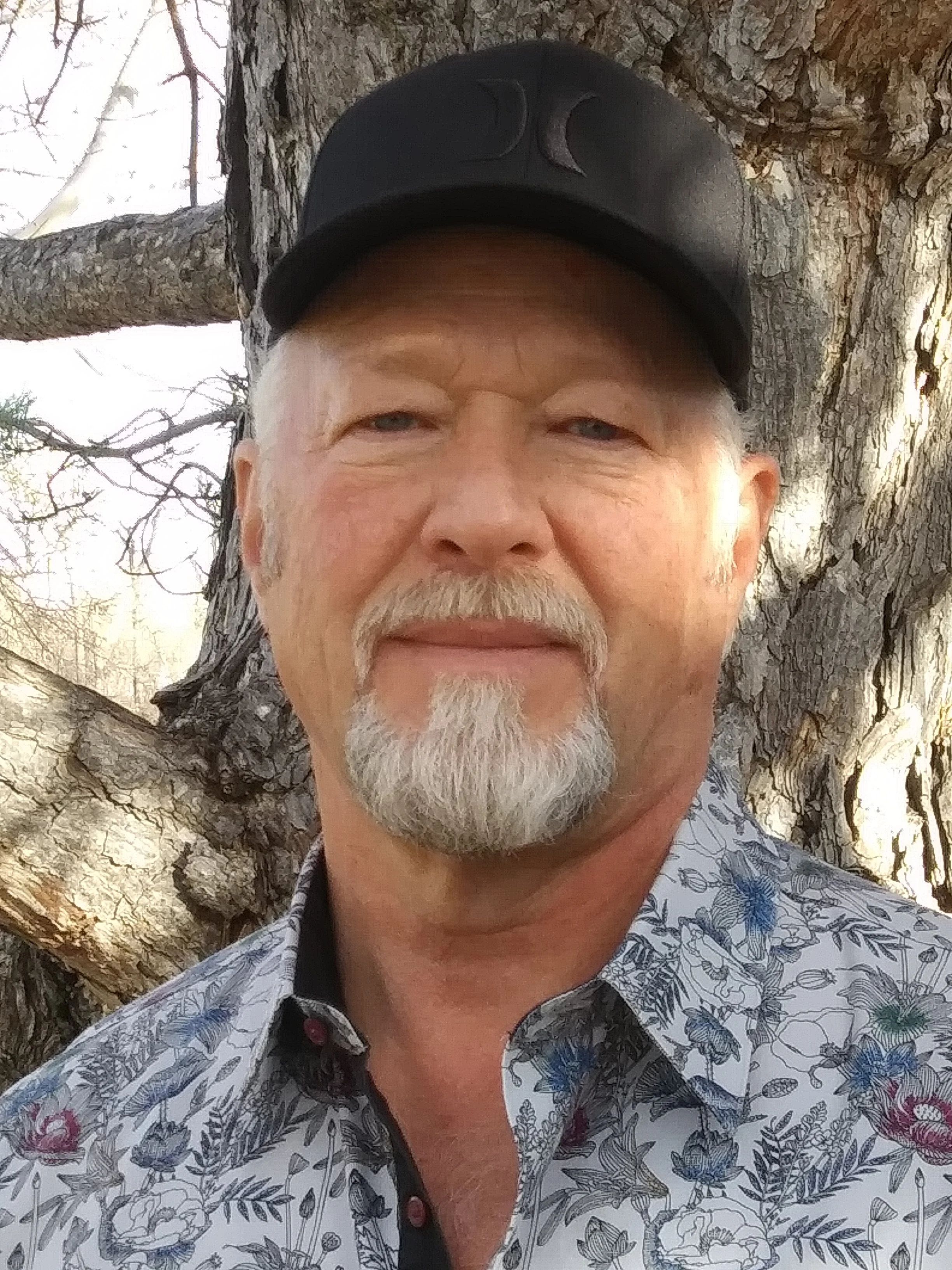 A man with a beard and mustache wearing a black cap with a moon symbol, standing in front of a large tree.