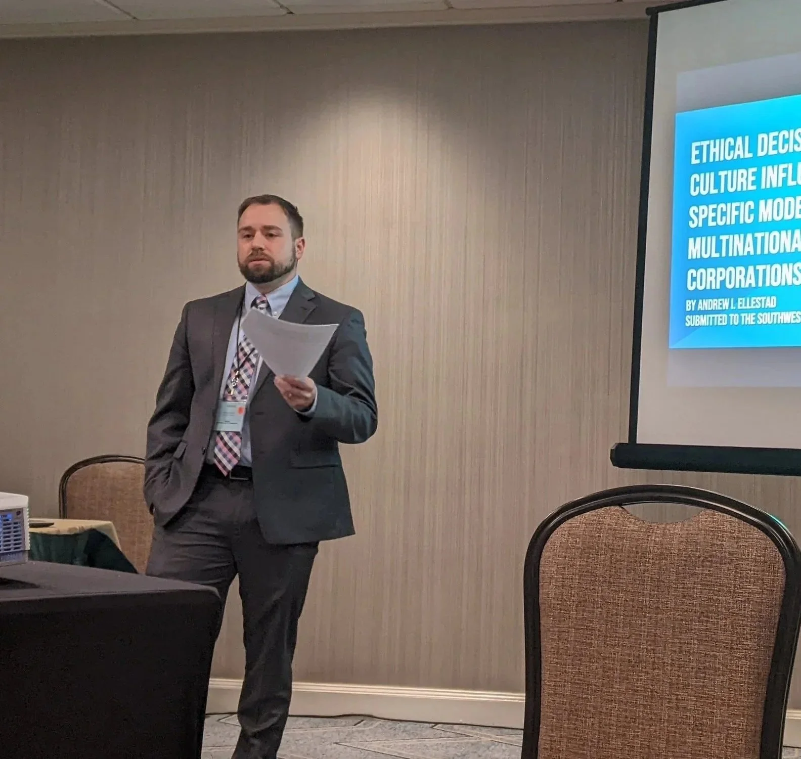 Man in a suit giving a presentation at a conference, holding papers, with a slide titled 'Ethical Decision-Making in Corporate Culture' projected on a screen to his right.
