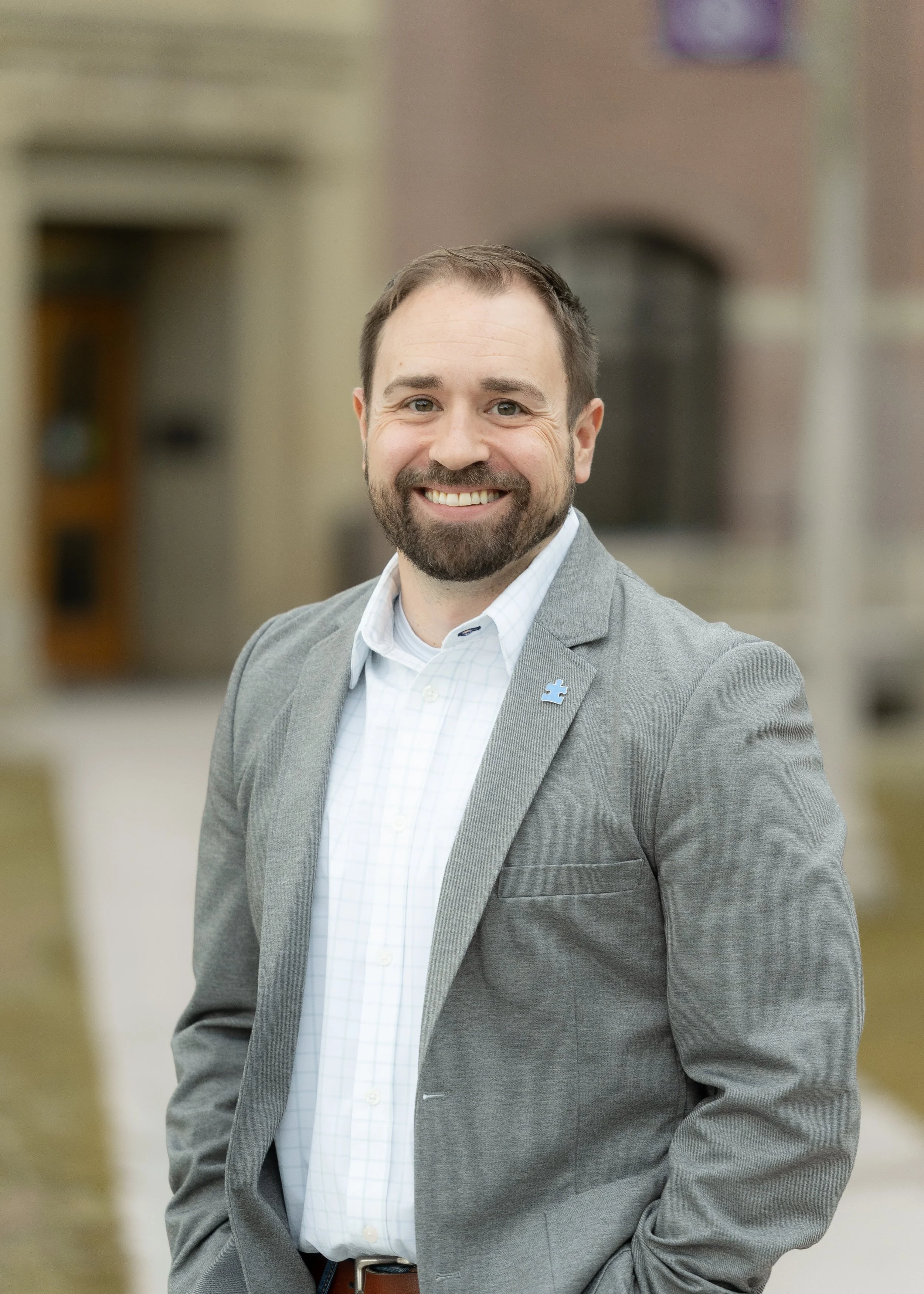 A smiling man in a gray blazer and white shirt standing outdoors with a building in the background.