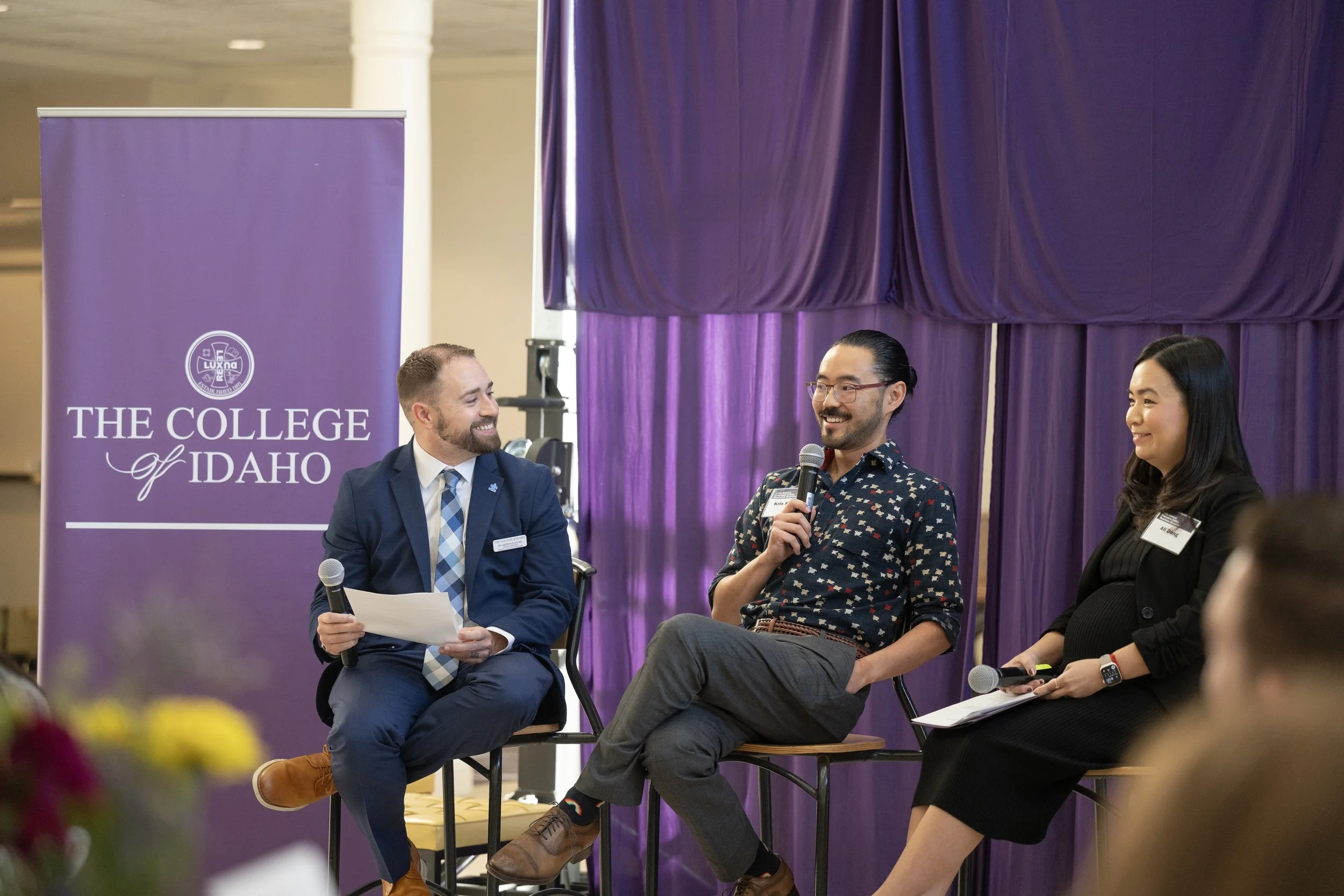 Three people seated on a stage, participating in a panel discussion or event. Behind them is a purple curtain and a purple banner that says 'The College of Idaho.' The man on the left is wearing a suit and tie, holding a paper. The man in the middle, speaking into a microphone, is wearing glasses and a dark patterned shirt.