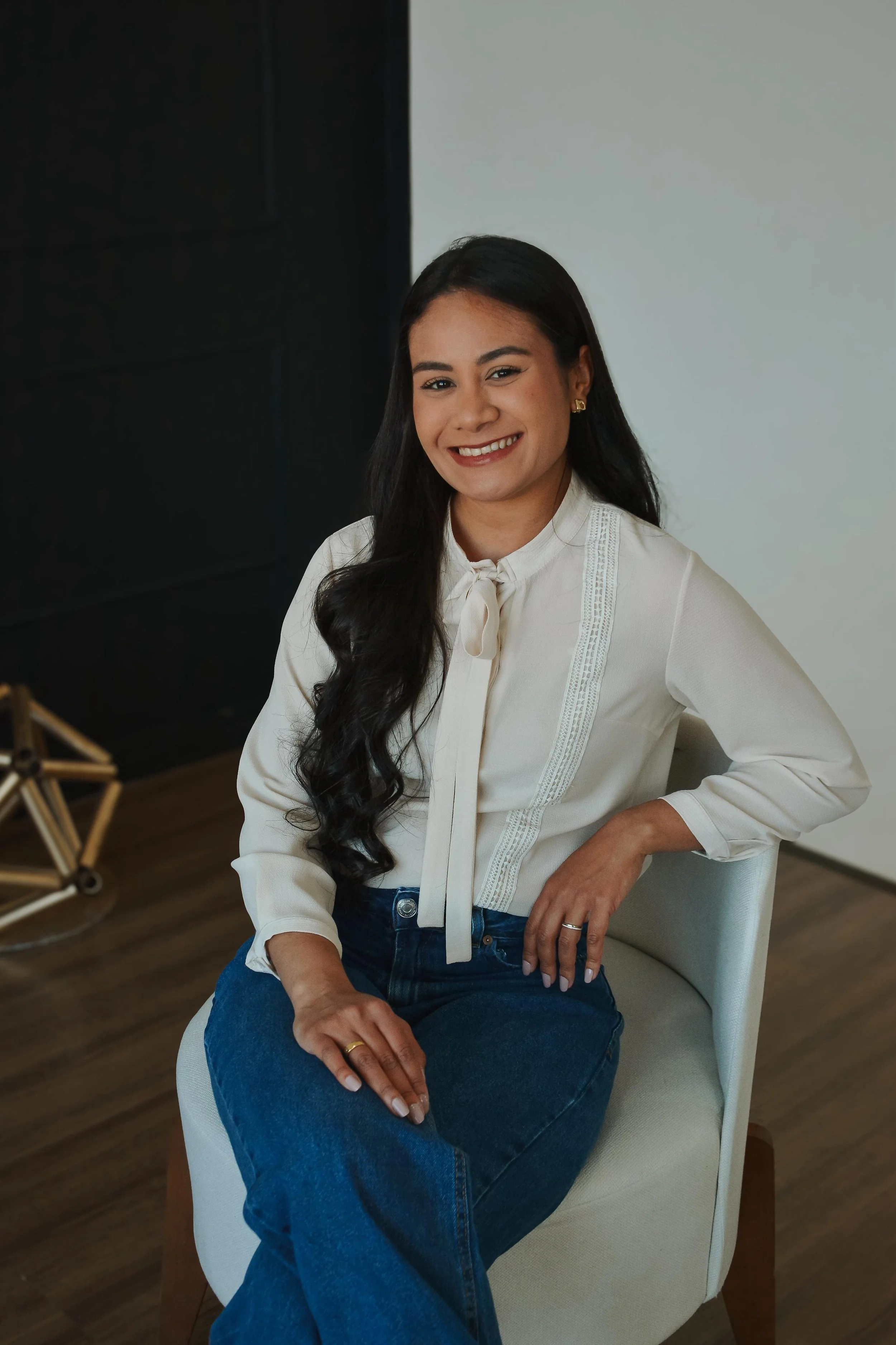 Mulher sorrindo, sentada em uma cadeira, usando uma blusa branca com laço no pescoço e jeans azul escuro, com cabelo longo e escuro, em um ambiente interno com piso de madeira e parede branca.