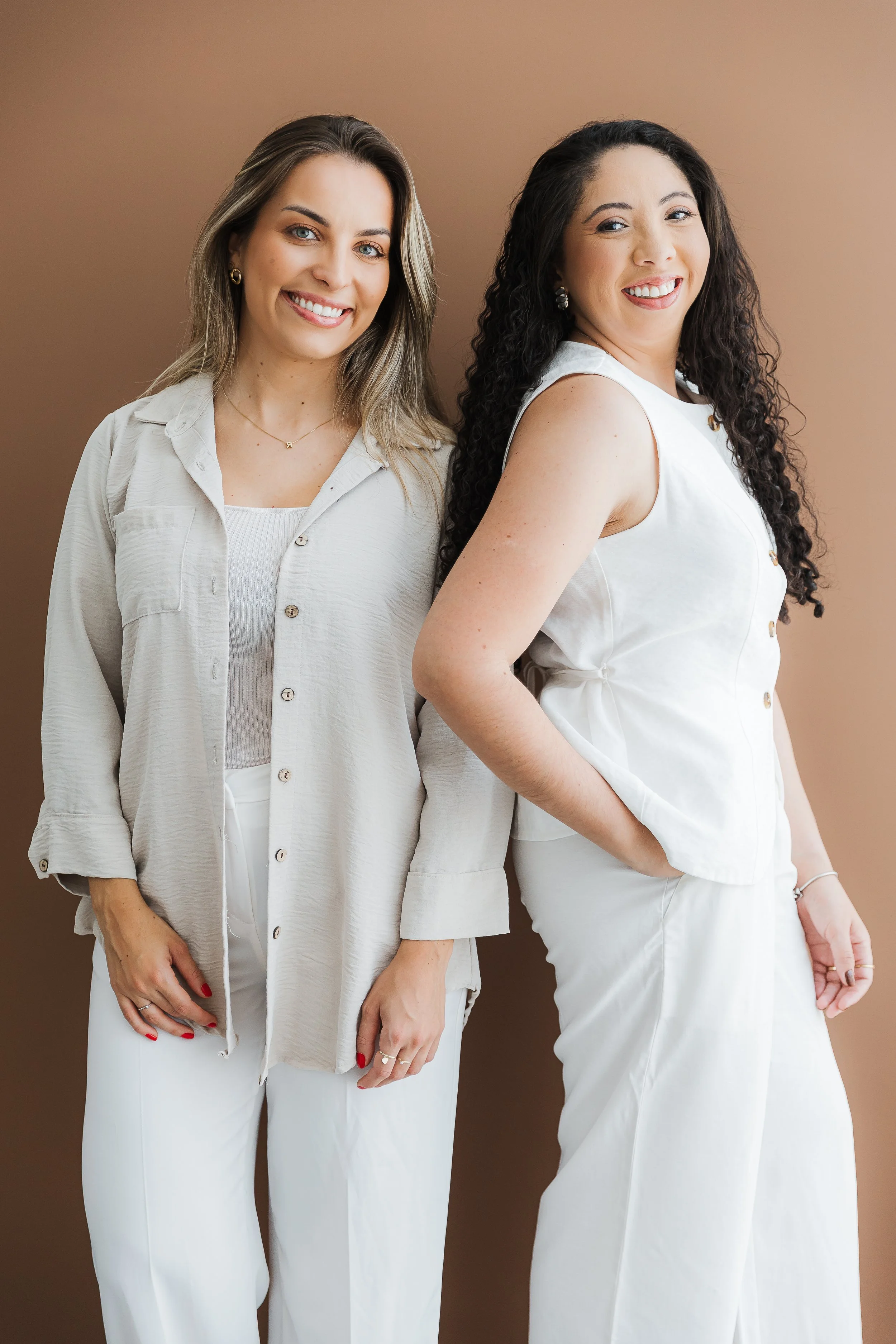 Duas mulheres sorrindo usando roupas brancas, uma com cabelo loiro e a outra com cabelo cacheado escuro, posando juntas contra fundo marrom.