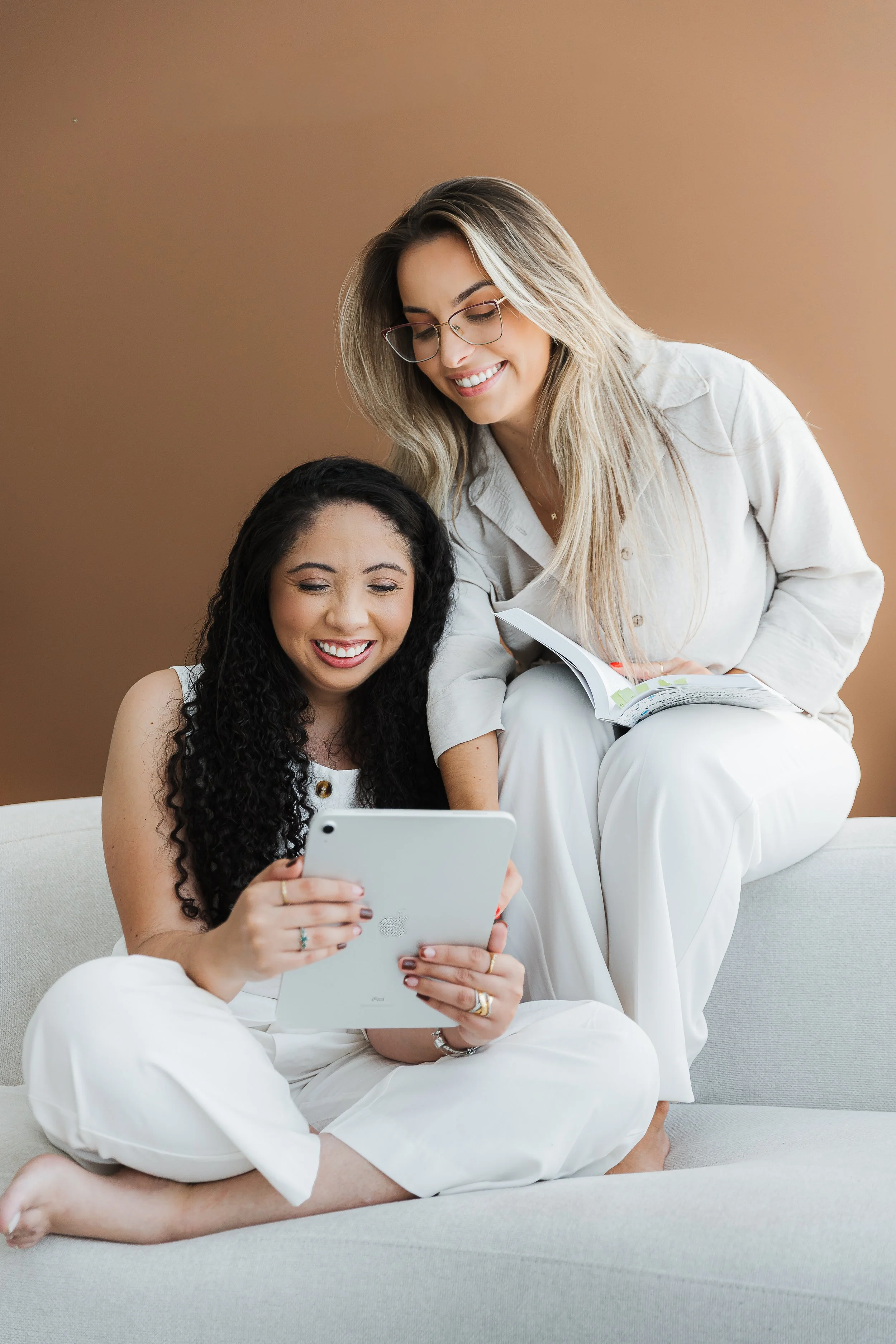 Duas mulheres, uma sentada no sofá segurando um tablet e outra sorridente, sentada no colo da primeira, lendo um livro.