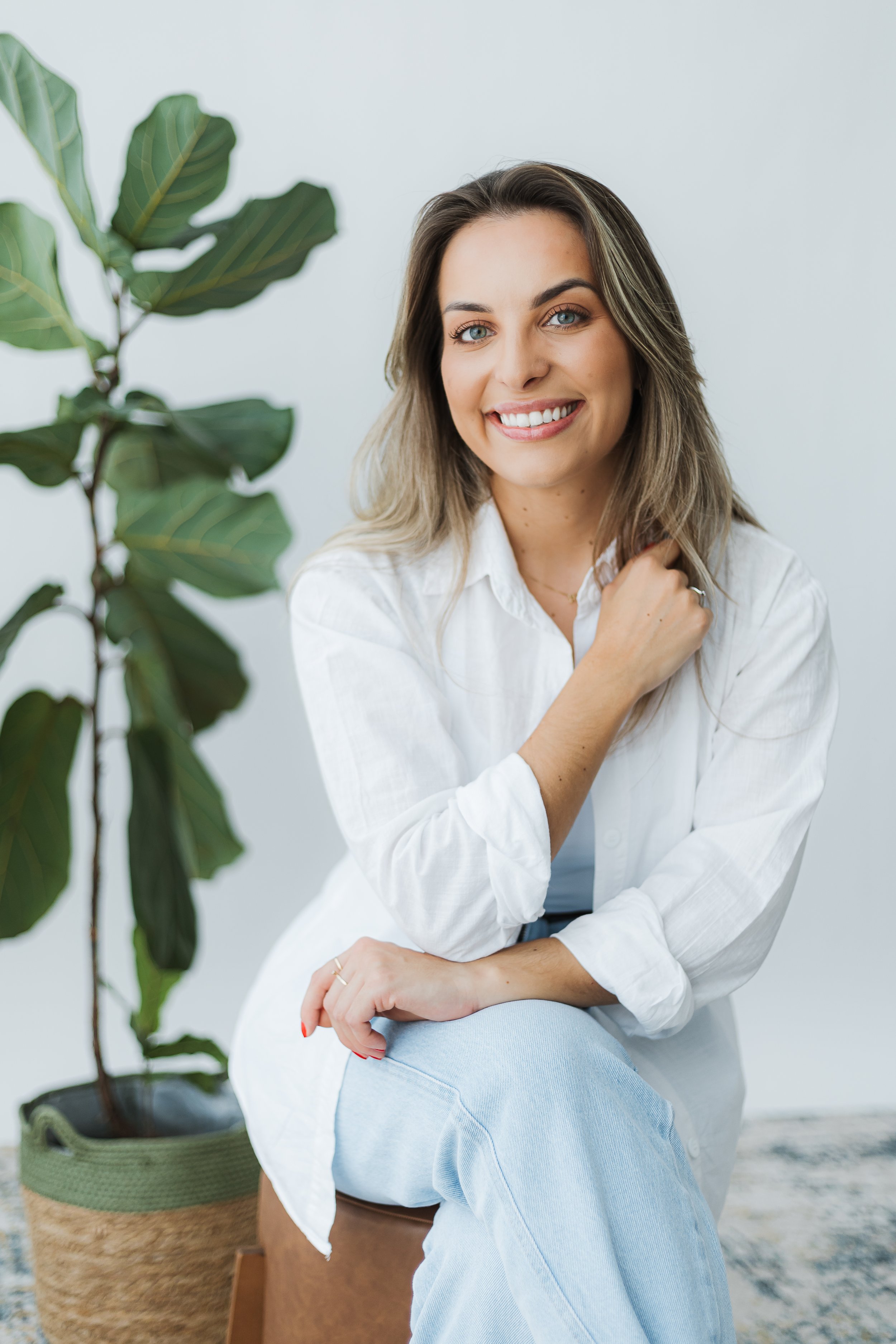 Mulher sorridente com cabelo castanho e olhos azuis, vestindo camisa branca, sentada ao lado de uma planta grande, com fundo branco.