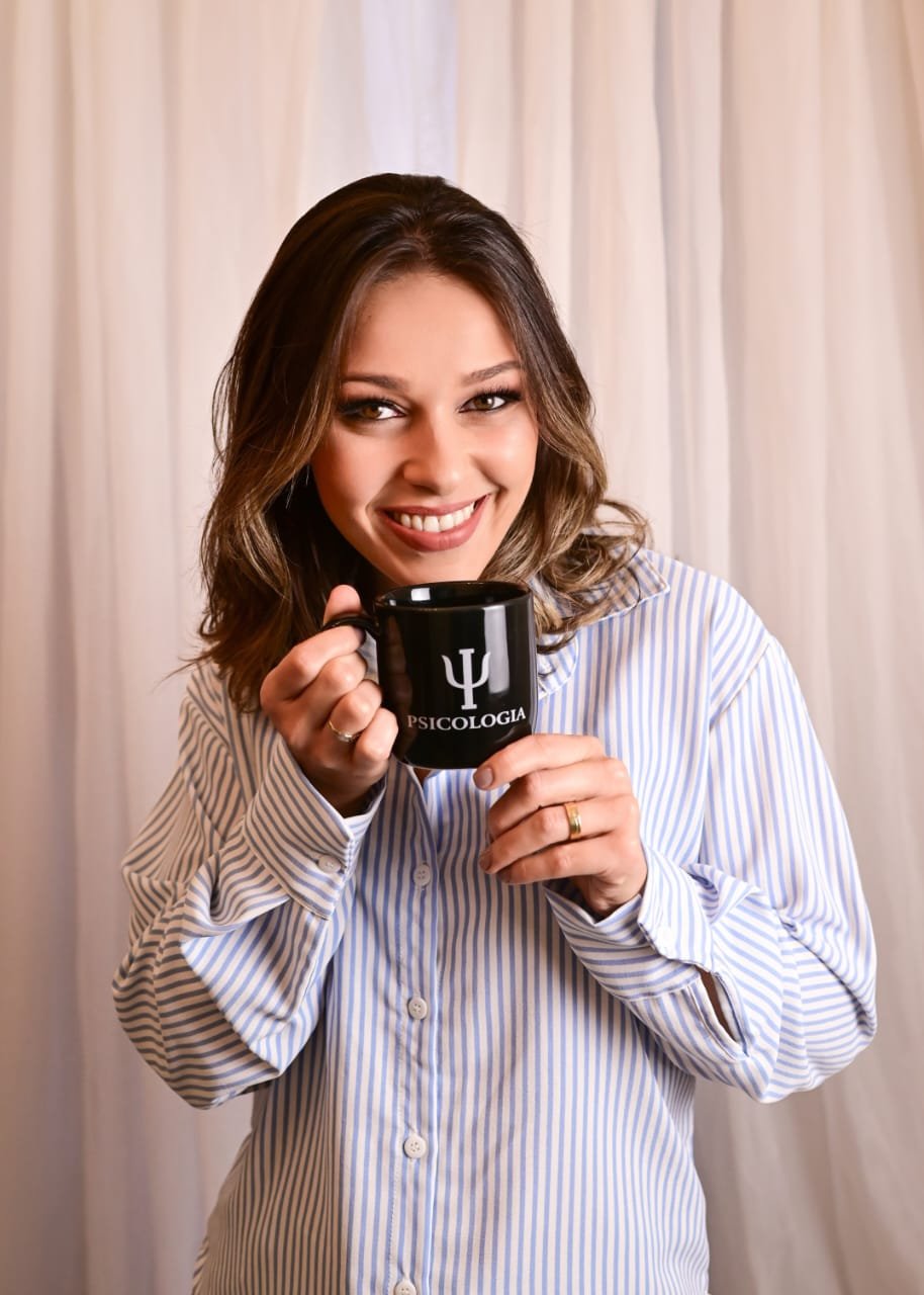 Mulher sorridente segurando uma caneca preta com o símbolo de psicologia e a palavra "Psicologia" escrito nela, em fundo de cortina branca.
