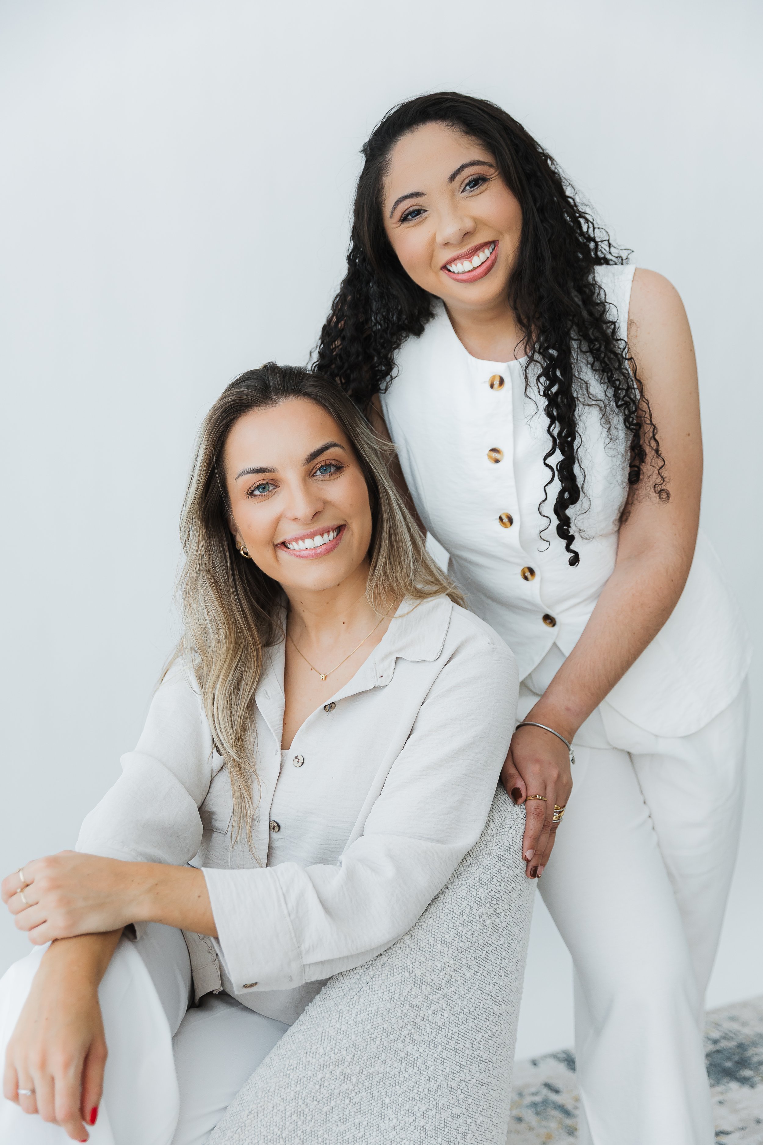 Duas mulheres sorridentes posando para uma foto em um ambiente branco, uma sentada e outra de pé atrás dela, ambas vestindo roupas brancas.