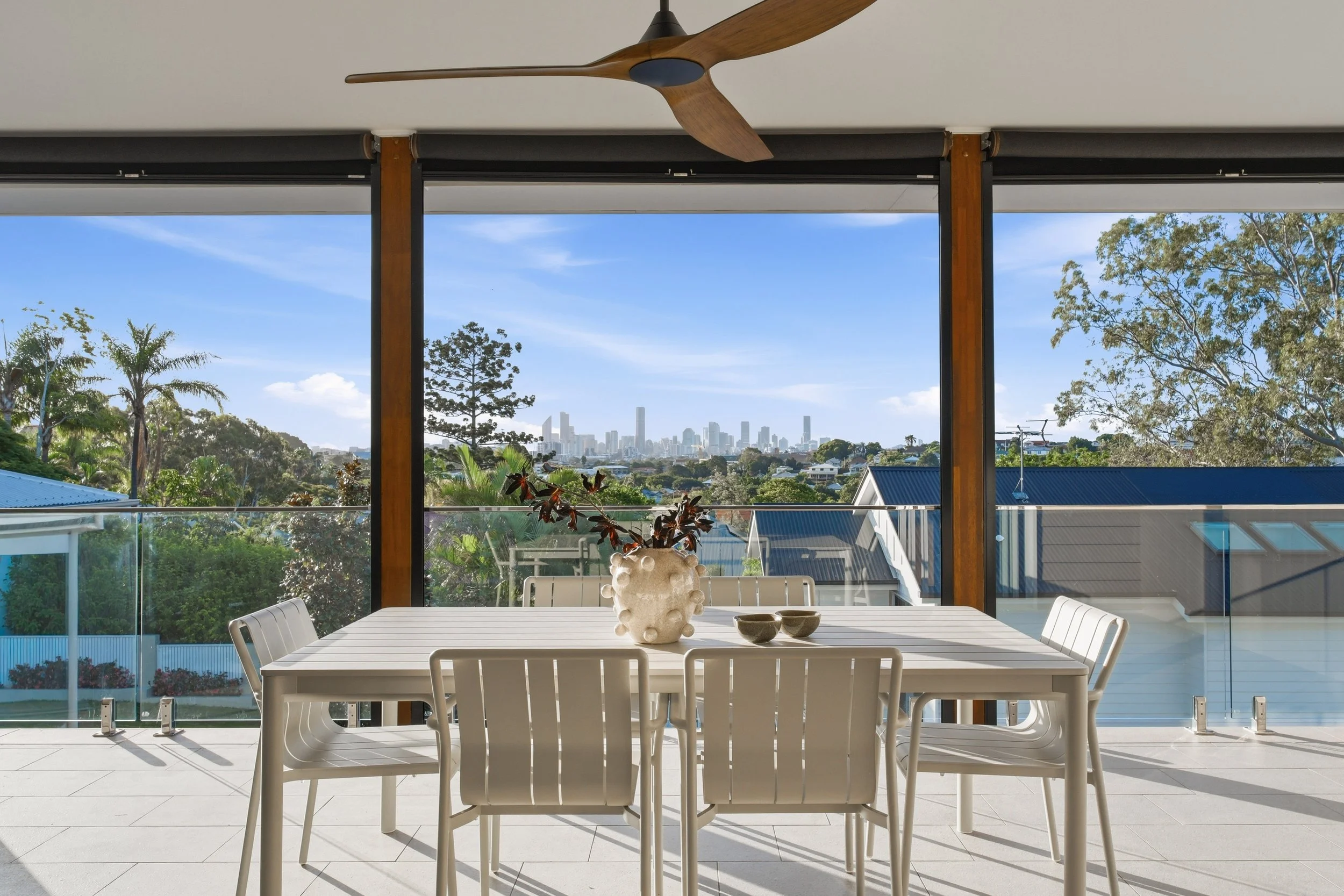 Modern outdoor dining area with a white table and chairs, decorative vase, and view of city skyline with tall buildings and trees under a blue sky