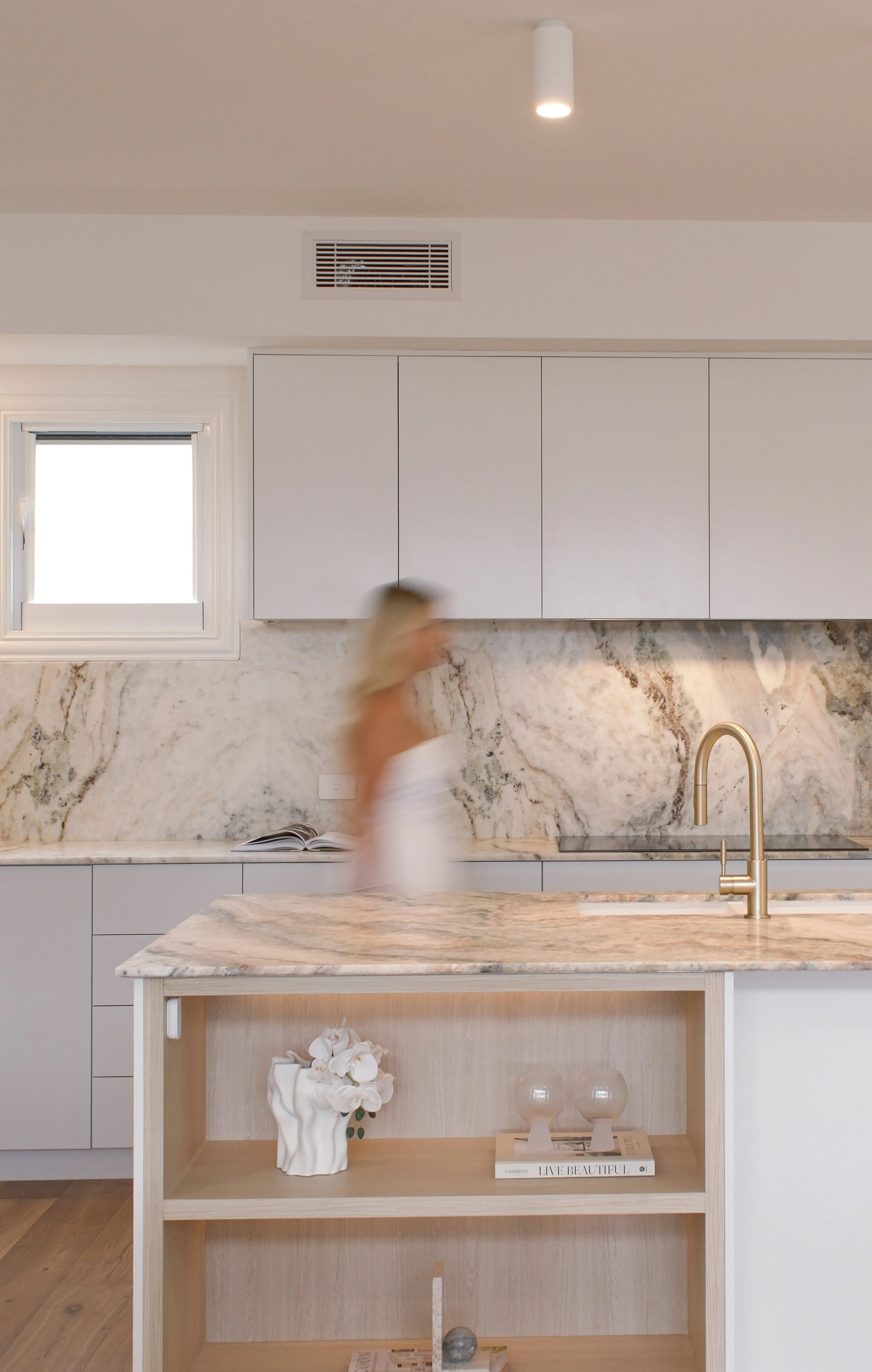 Modern kitchen with marble countertops, white cabinetry, and a kitchen island with open shelving. There's a small window above the countertops, a gold faucet, and decorative items including a white flower vase, books, and small sculptures.