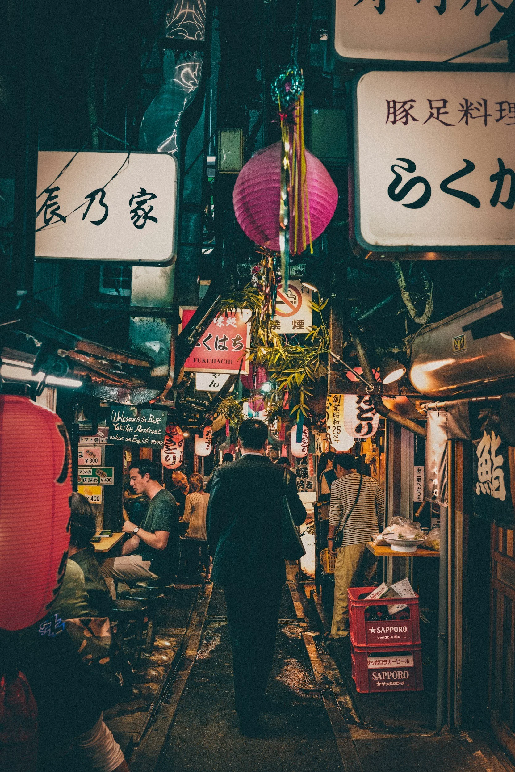 People walking through a narrow, vibrant Japanese street lined with signs, lanterns, and food stalls at night.