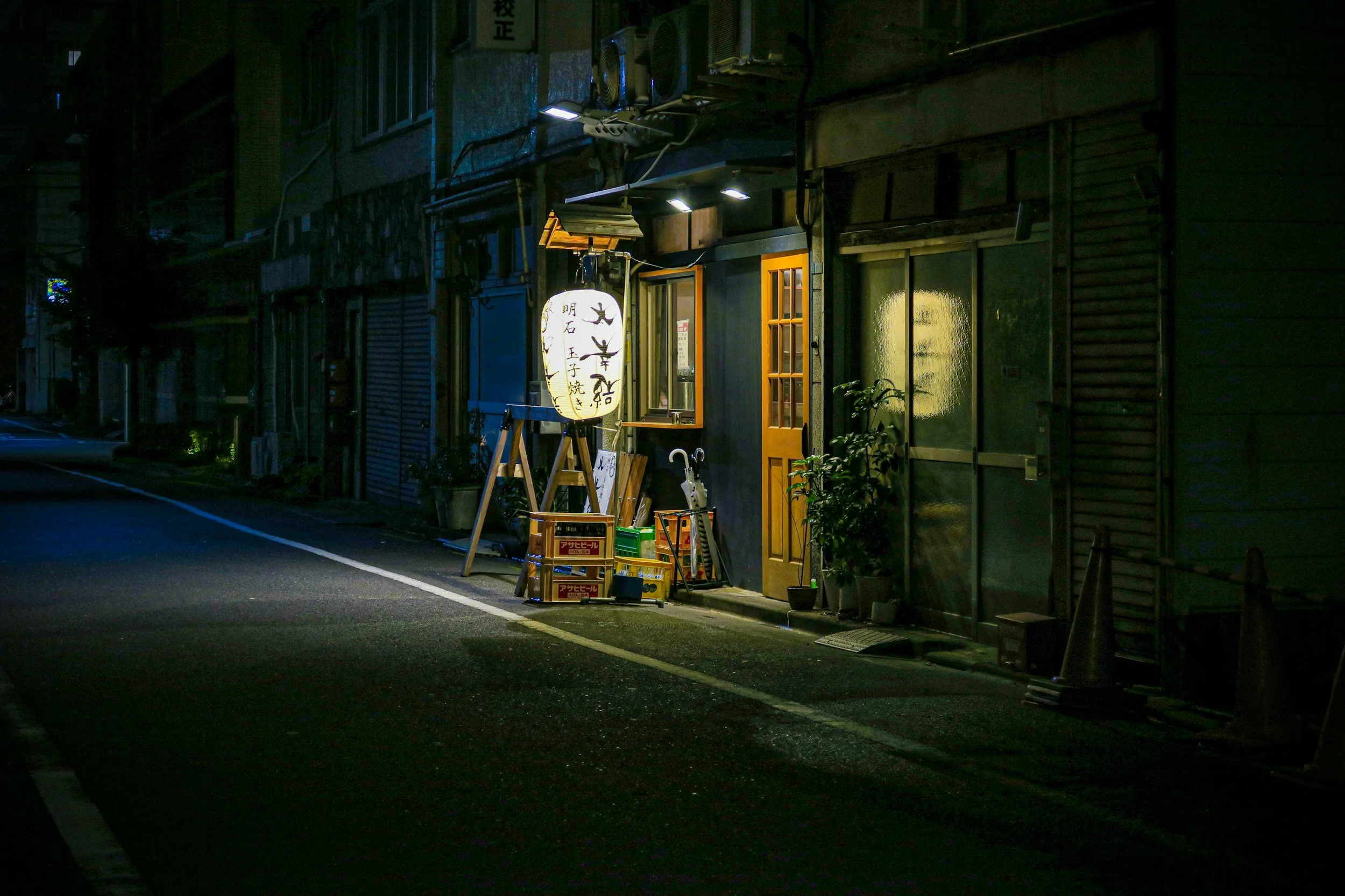 A dimly lit narrow street scene outside a small restaurant or shop, with a glowing paper lantern, some potted plants, an open window, and various boxes and umbrellas near the entrance.