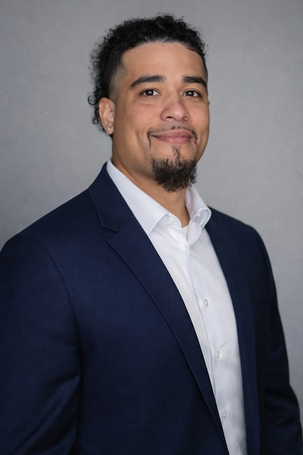 A man in a dark blue suit and white shirt smiling against a gray background.