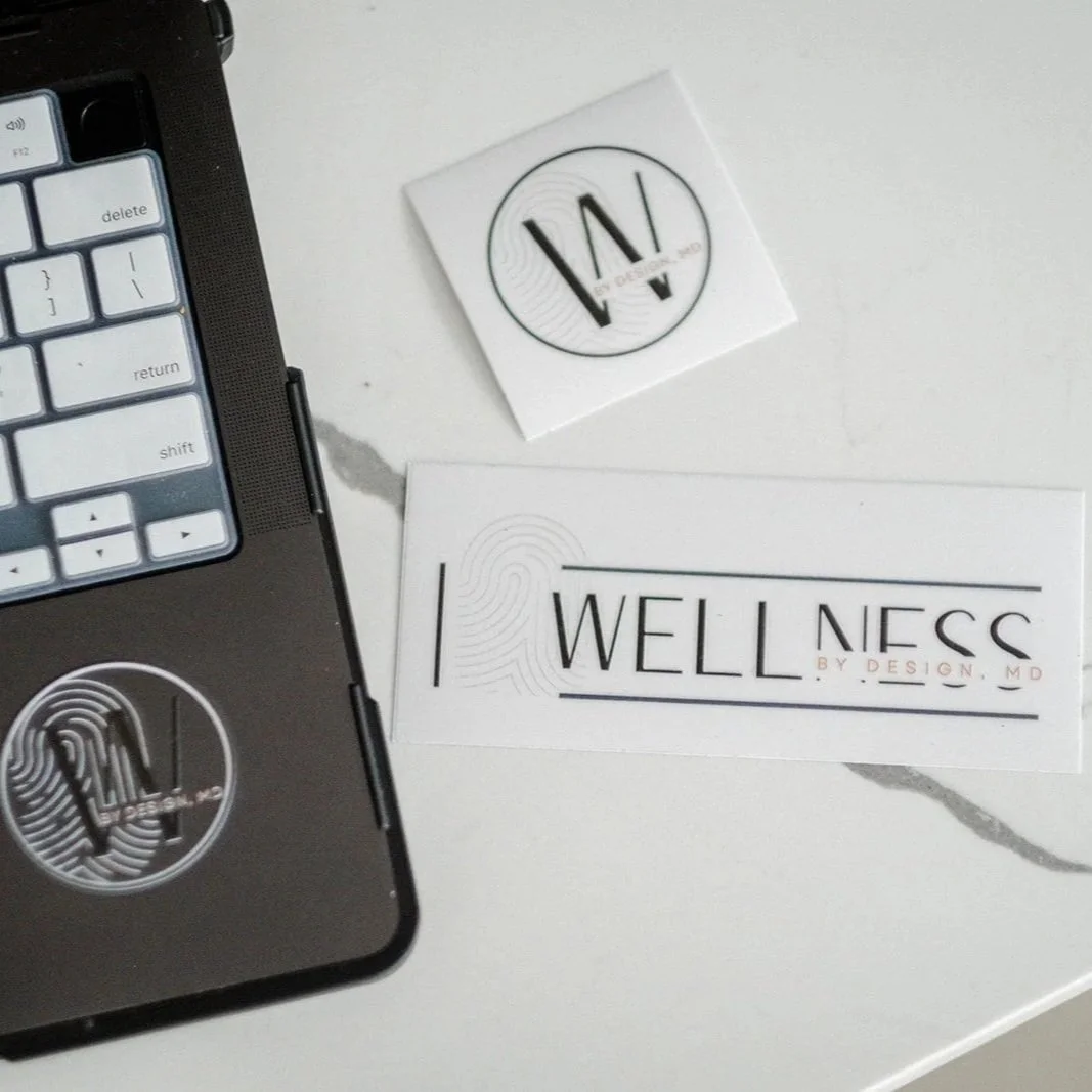 A close-up view of a laptop keyboard and two business cards on a white surface, featuring the logos of 'WELLNESS BY DESIGN, MD' and a circular logo with headphones.