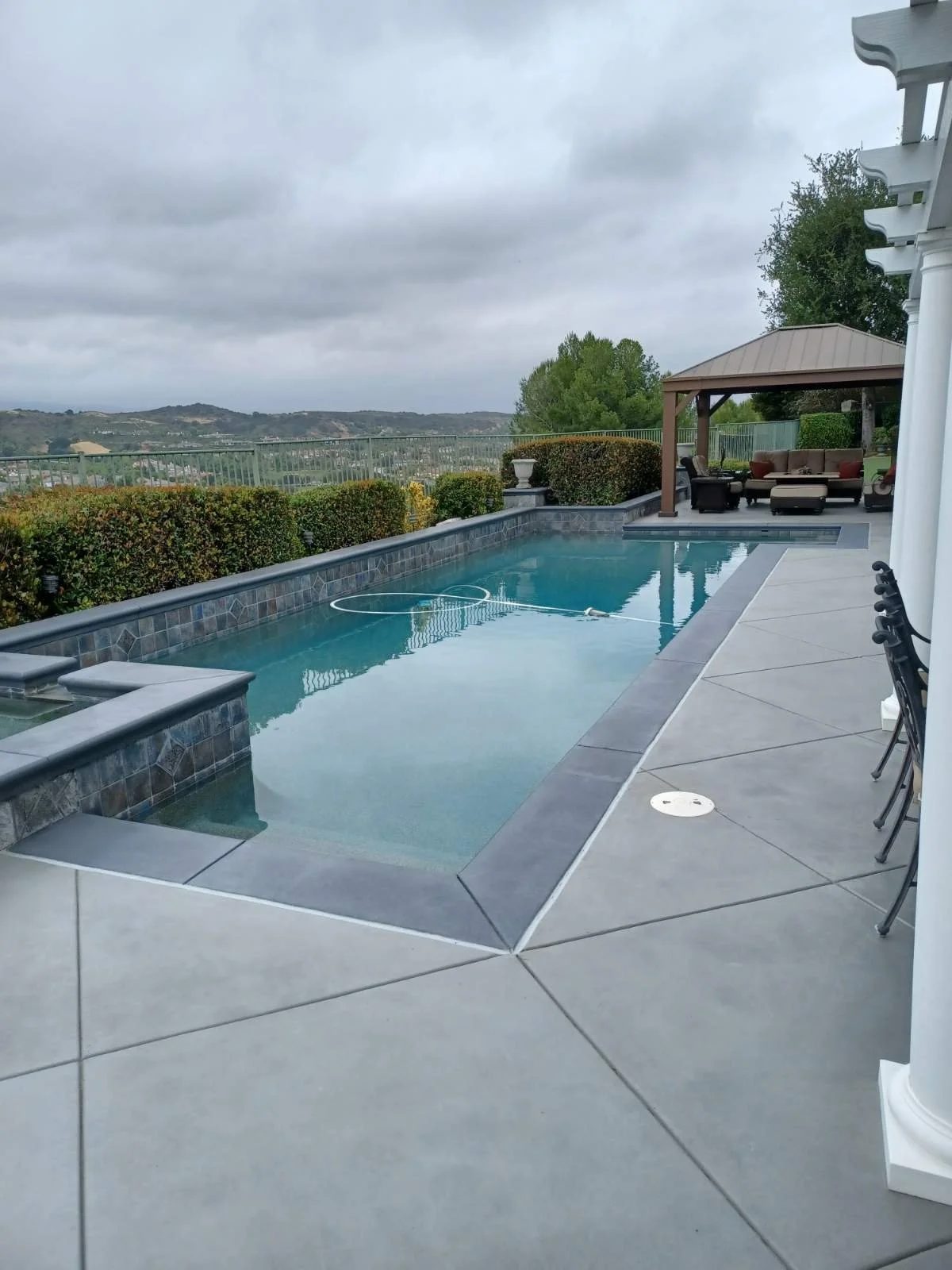 Empty backyard swimming pool with a spa, surrounded by concrete and a seating area with outdoor furniture, under a cloudy sky.