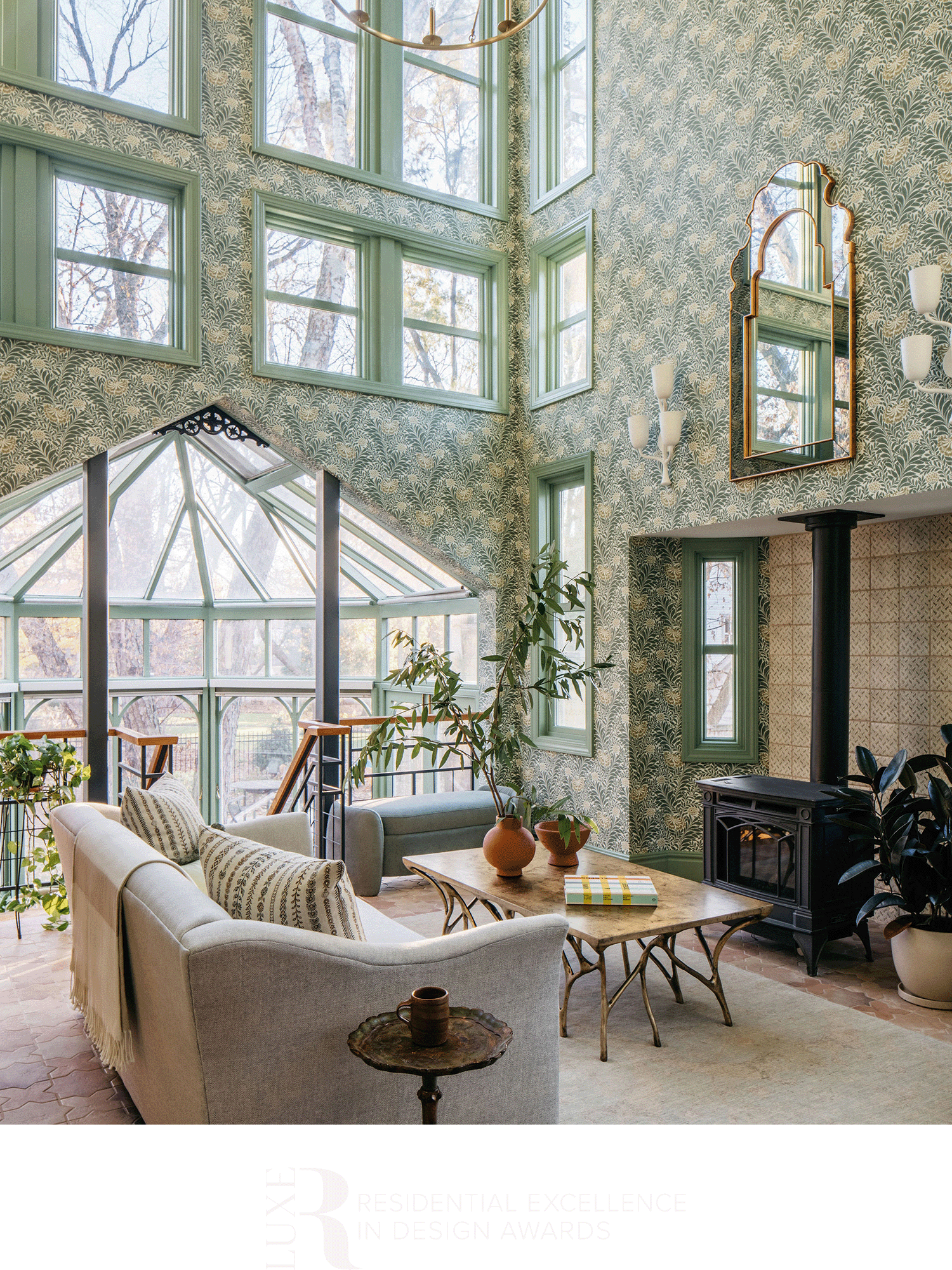 A cozy living room with large windows, a patterned wallpaper, and a mix of modern and vintage furniture, including a sofa, armchair, coffee table, and a wood-burning stove.