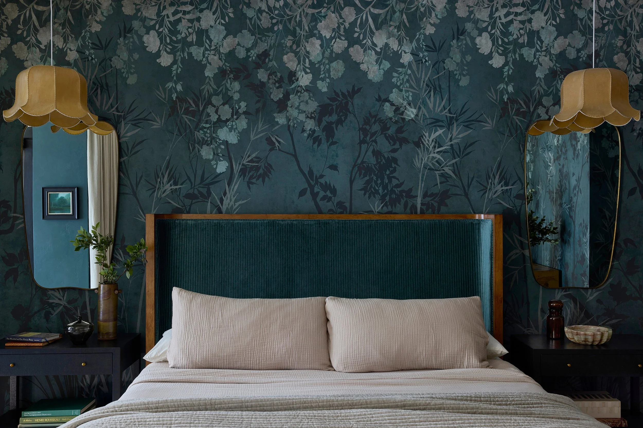 A bedroom with dark leafy wallpaper, a teal upholstered headboard, beige bedding, and two hanging mirrors with yellow lampshades.