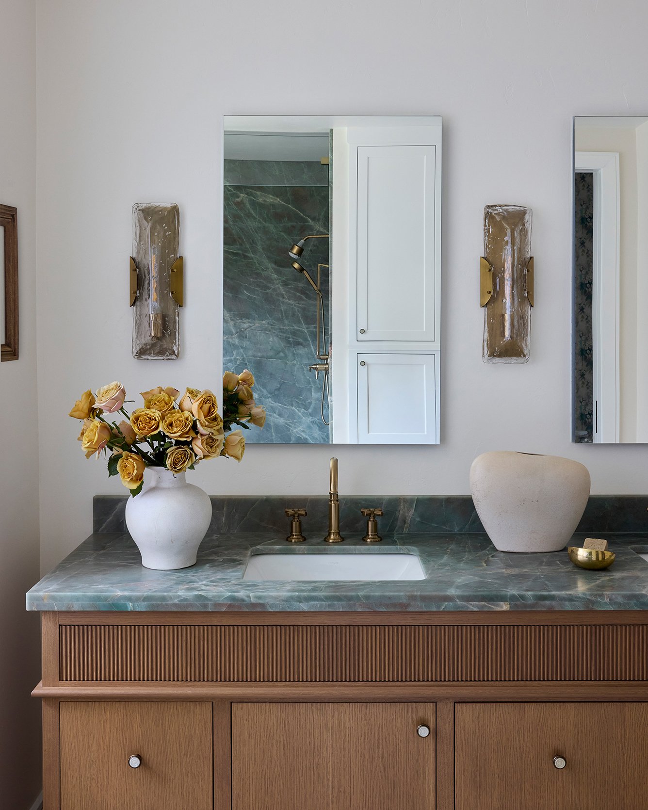 Bathroom vanity with a marble countertop, a white vase with yellow roses, and a beige decorative object, reflected in a mirror with wall-mounted lights on either side.