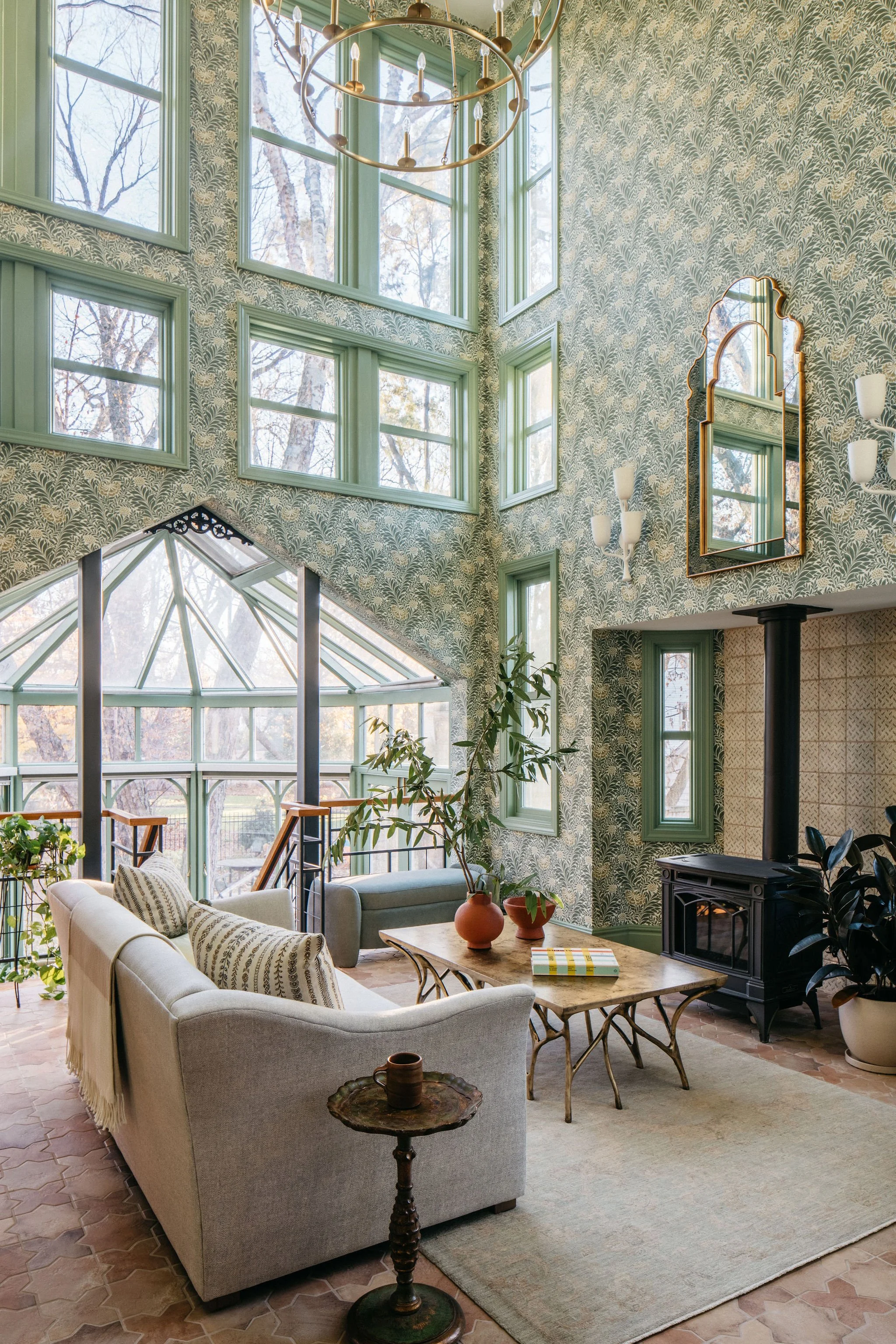 A spacious living room with tall, multi-paned windows and green framed walls, decorated with botanical wallpaper. There is a round chandelier hanging from the ceiling, a large mirror with a gold frame, a wood-burning stove, a sofa, armchairs, a coffee table with vases and books, and various indoor plants.