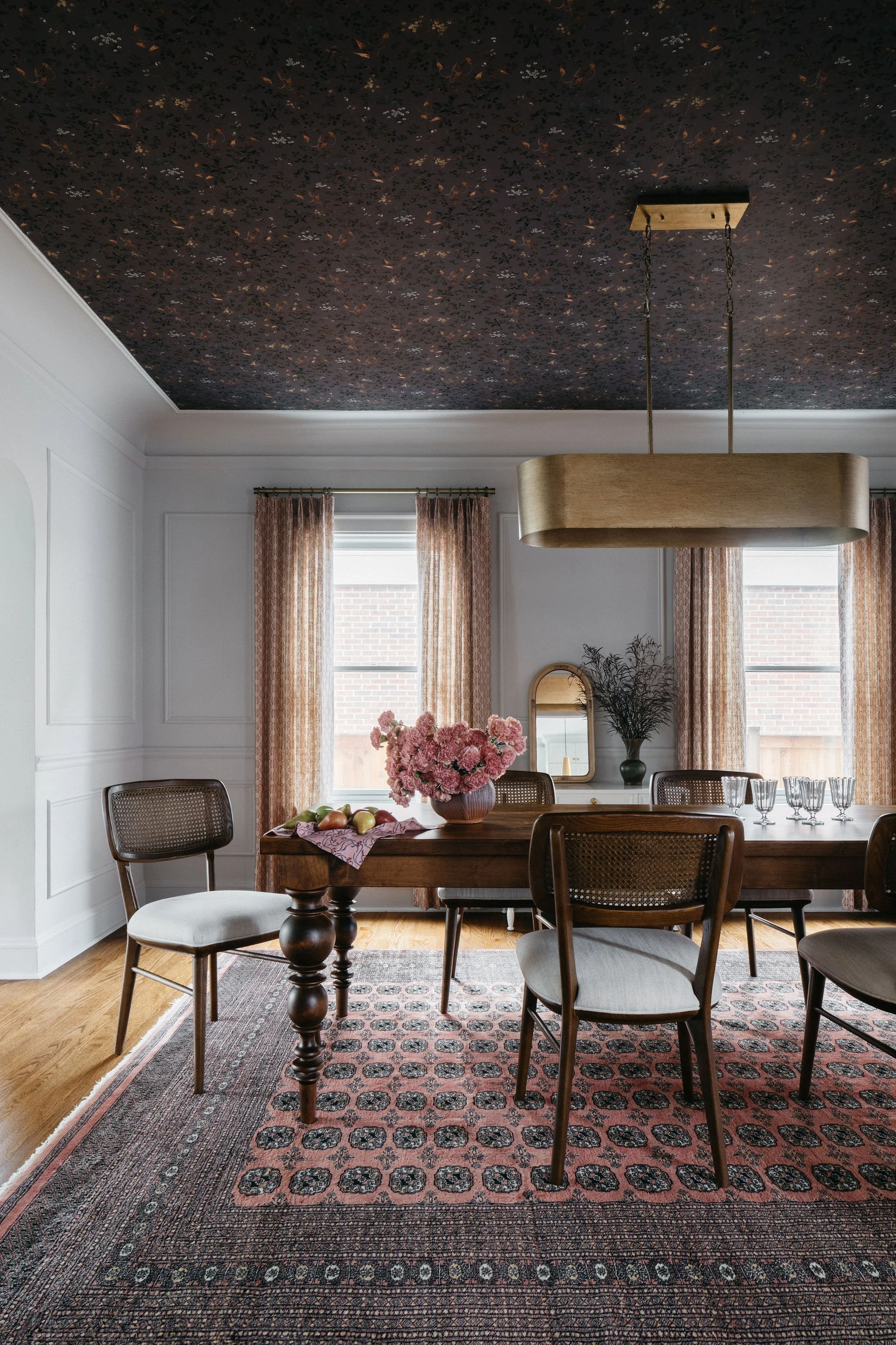 A dining room with a wooden table surrounded by six chairs; a pink flower centerpiece, a few apples, and glasses are on the table. Two windows with patterned curtains, a mirror, and a vase with branches are in the background, and a large rectangular light fixture hangs from the dark, star-patterned ceiling.