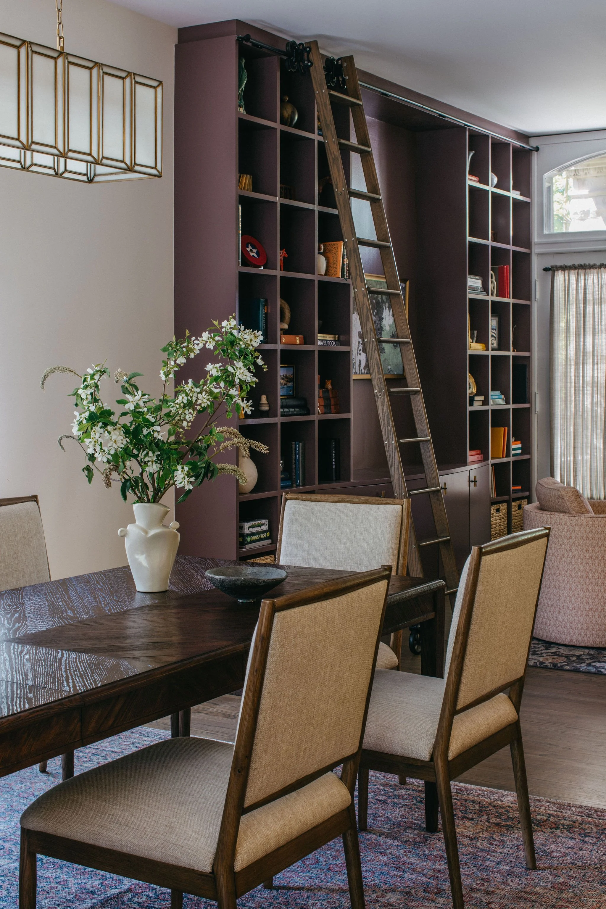 Dining room with a dark wood table, beige upholstered chairs, a vase with white flowers, and a large built-in bookshelf with a ladder and decorative items.