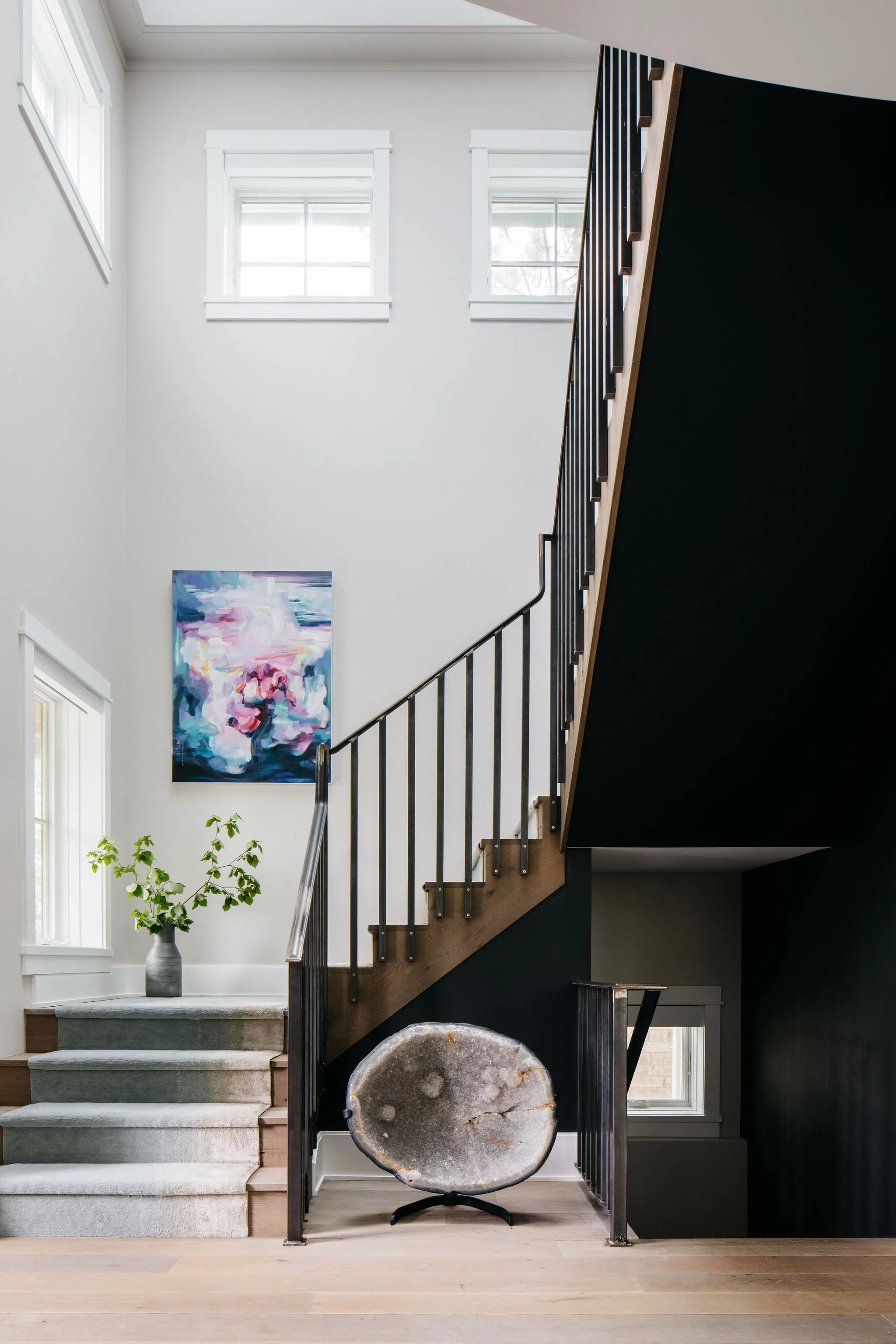 Interior of a modern home staircase area featuring white walls, two small upper windows, a contemporary painting, a potted plant, and a decorative stone or crystal sculpture on a stand.
