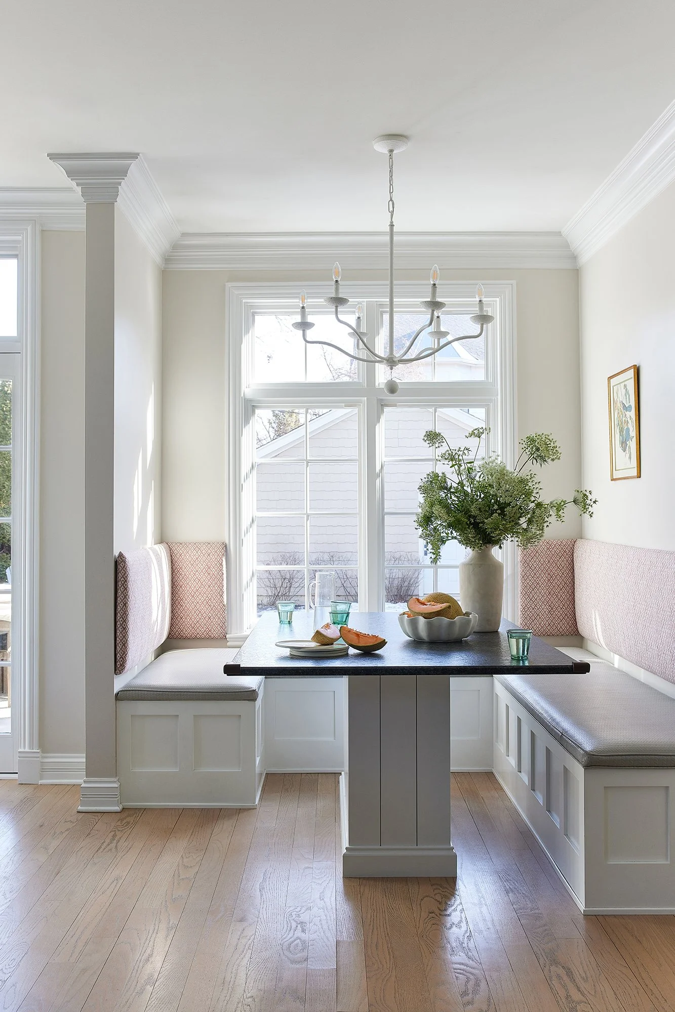 Bright corner breakfast nook with built-in seating, a black table, a large white vase with green foliage, and a window with views of neighboring houses.