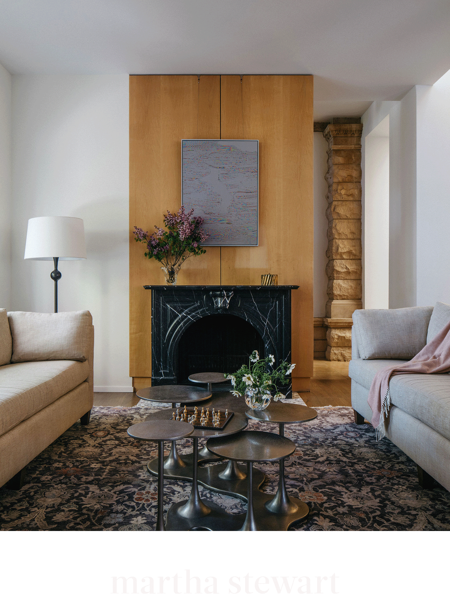 Living room with a black marble fireplace, beige sofas, a multi-tiered black metal coffee table, a white floor lamp, a vase with purple flowers, and a painting above the fireplace.