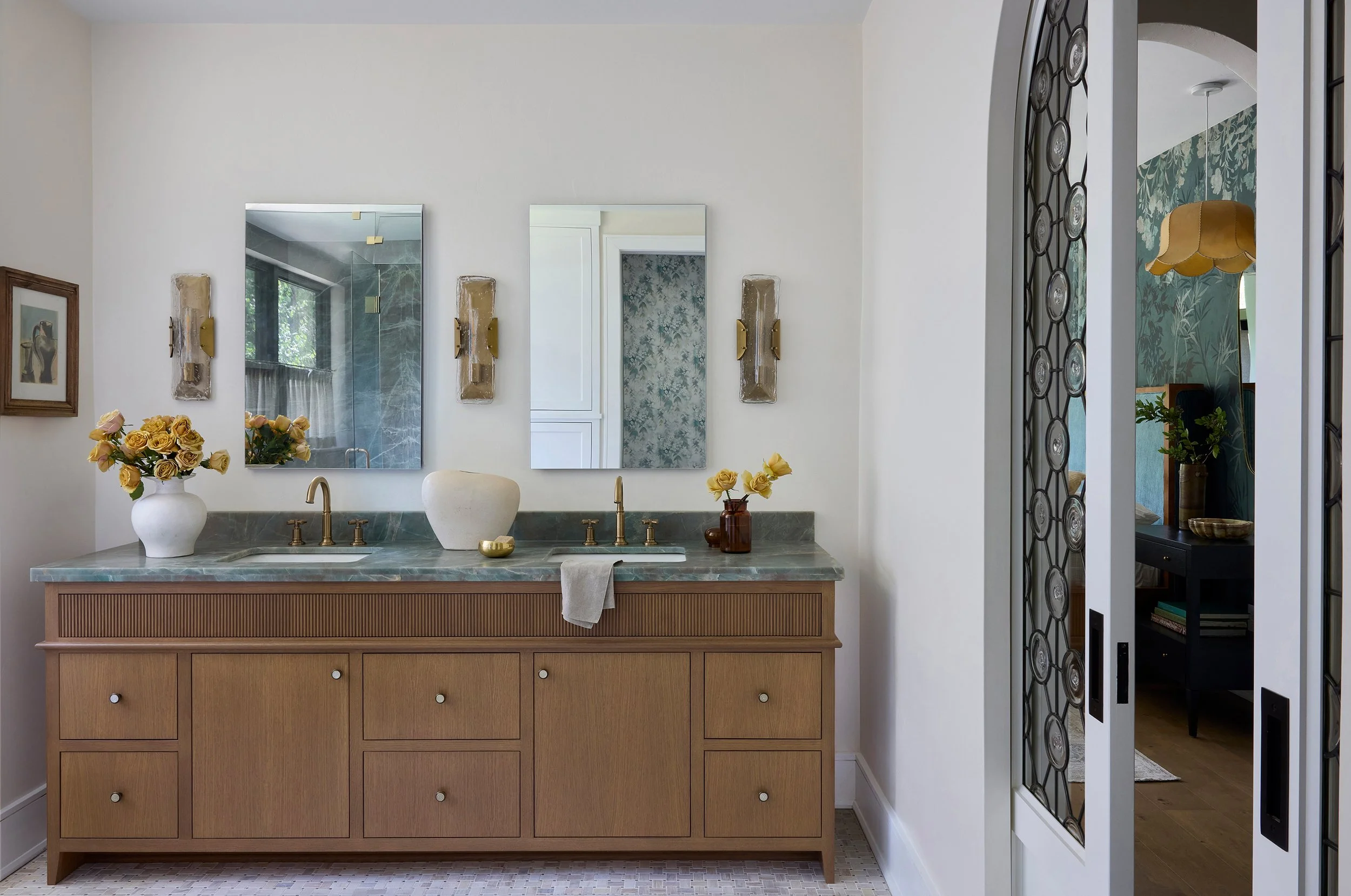 A bathroom with a wooden vanity and marble countertop, two mirrors above, vases with yellow roses, and a towel. Visible doorway with patterned wallpaper leading to another room with a dark side table and a lamp.