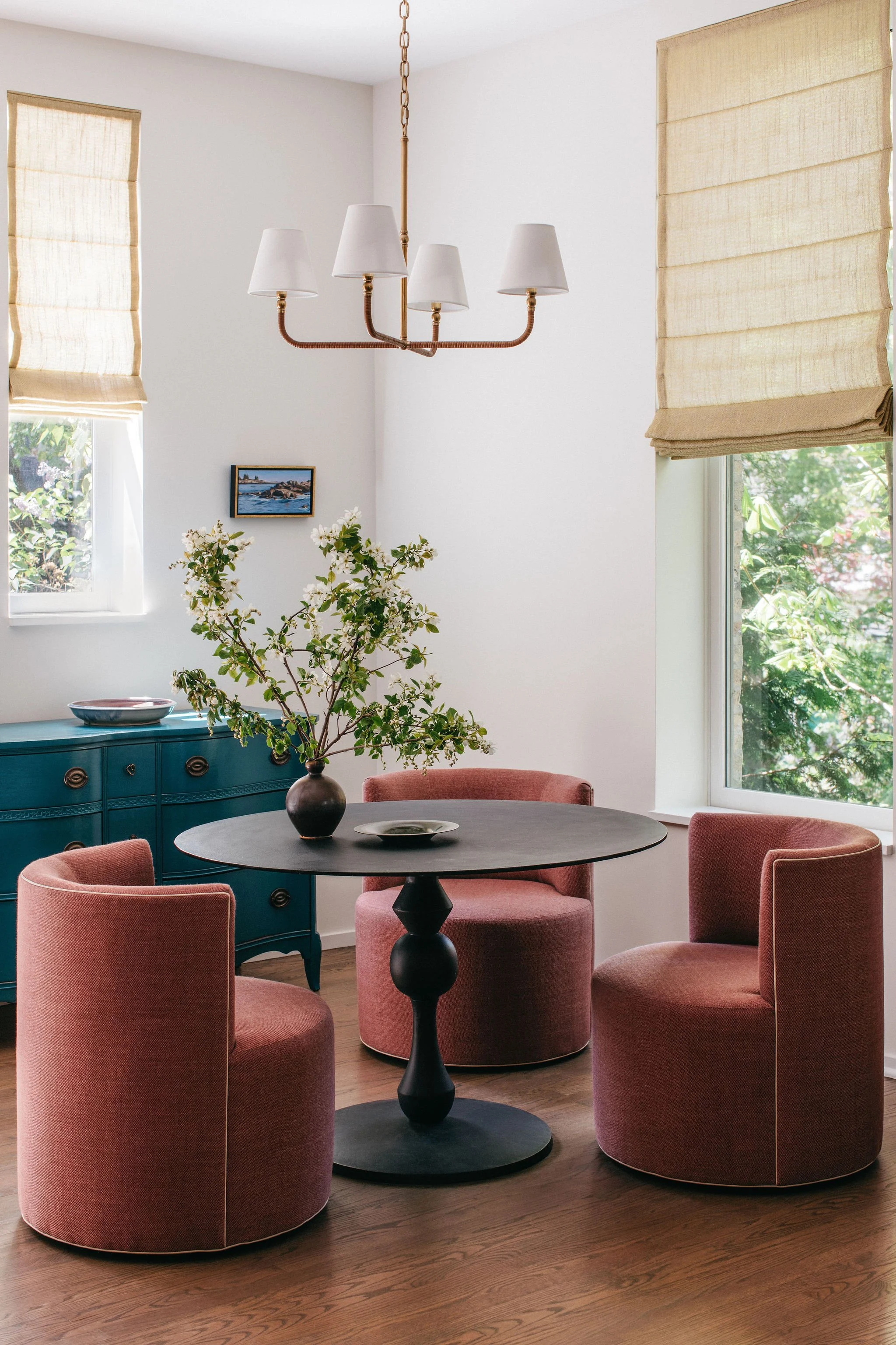 A dining room with a round black table, three pink upholstered chairs, a teal sideboard, a vase with white flowers, a wall hanging, large windows with beige blinds, and a chandelier with five white shades.