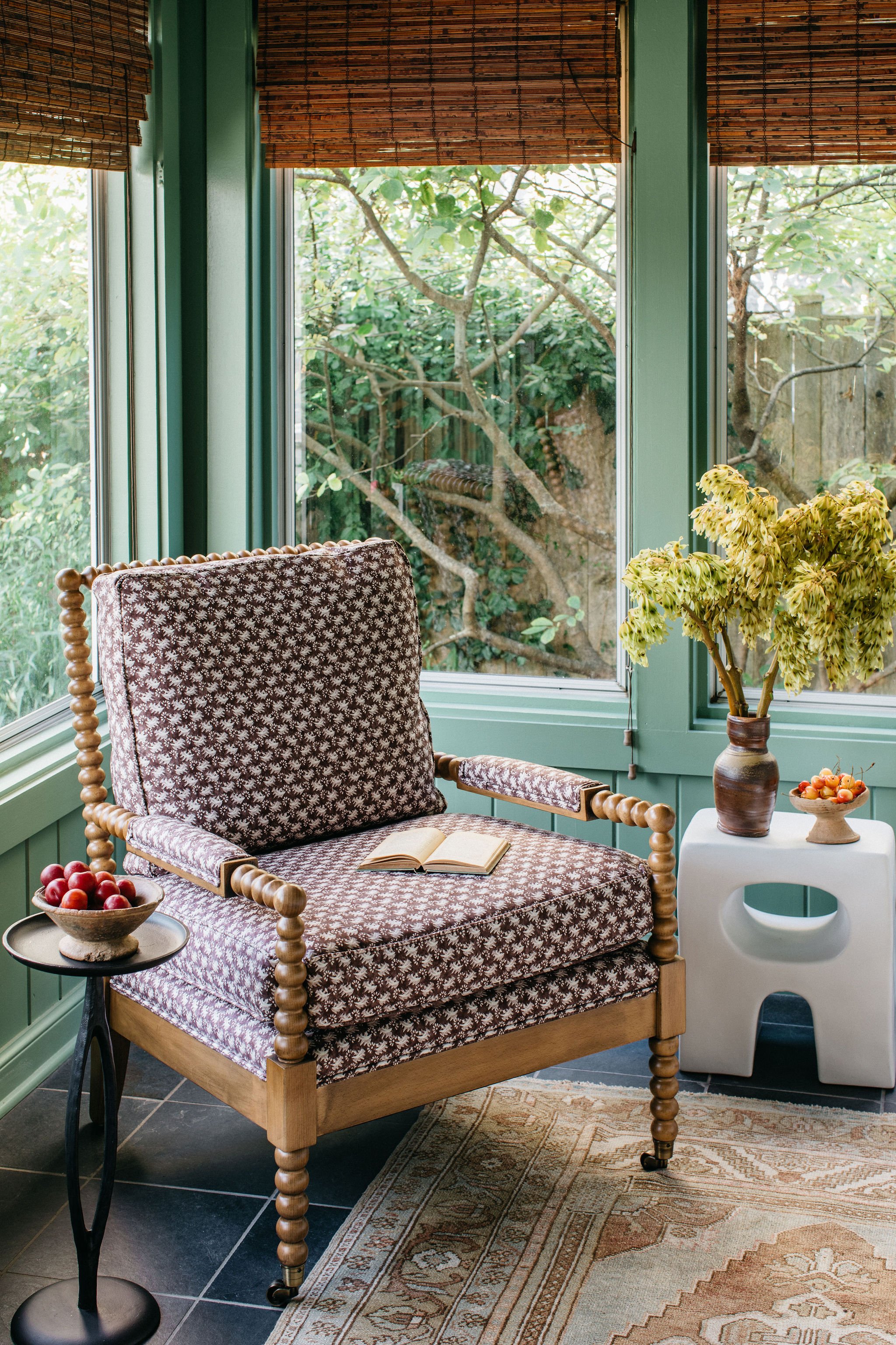 A cozy sitting area with a patterned armchair, a side table with apples, a white stool with a ceramic vase of dried flowers, and another bowl of small fruit, in front of large windows with green frames and bamboo blinds, overlooking a garden with trees and greenery.